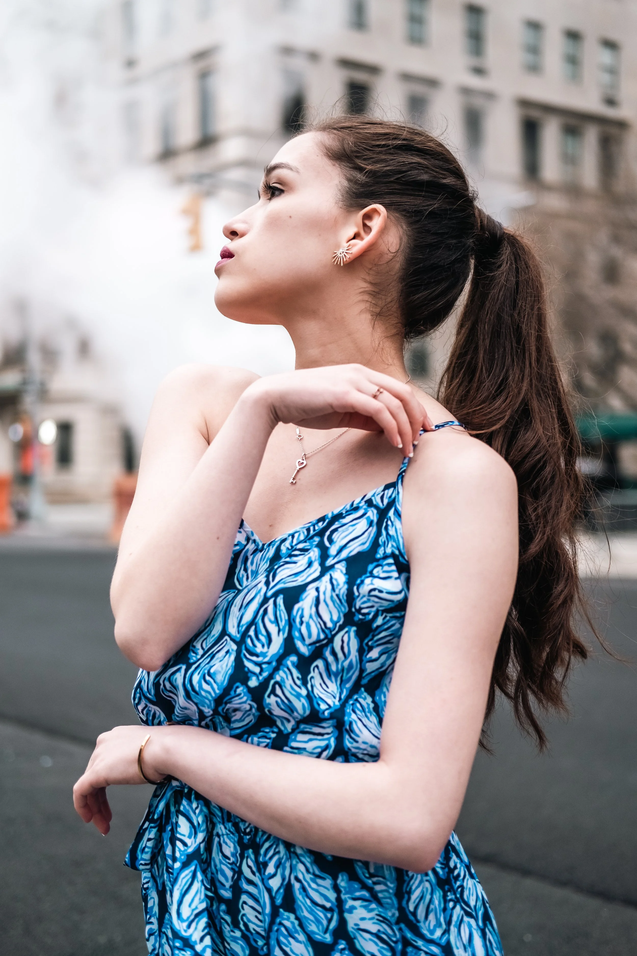 Side profile of a young woman with long brown hair wearing a blue and white patterned dress, earrings, necklace, and rings, standing outdoors in an urban environment with buildings in the background.