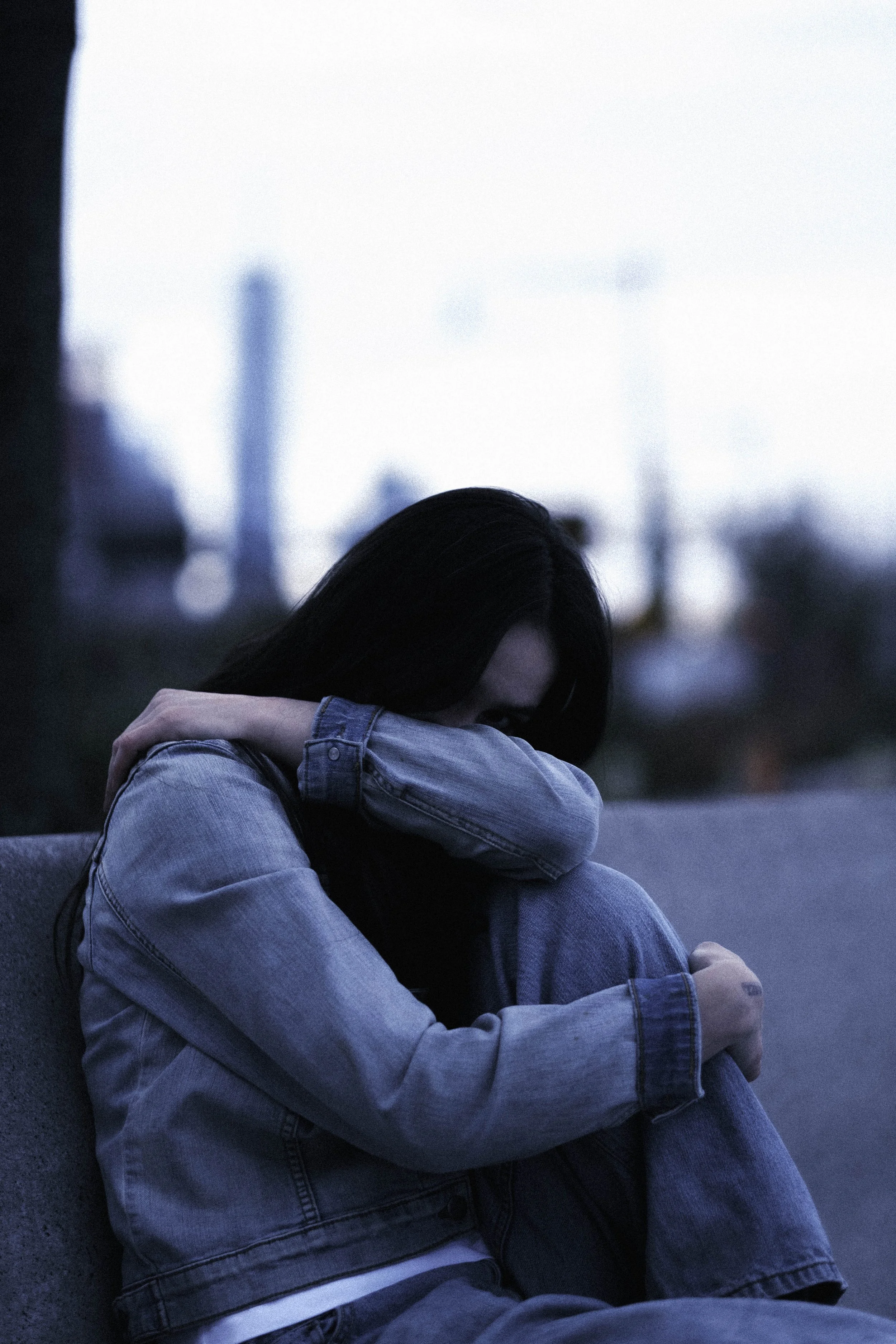A woman sitting on the ground with her knees pulled up and arms wrapped around them, wearing a denim jacket and jeans, with her head resting on her arm, in an urban setting during dusk or dawn.