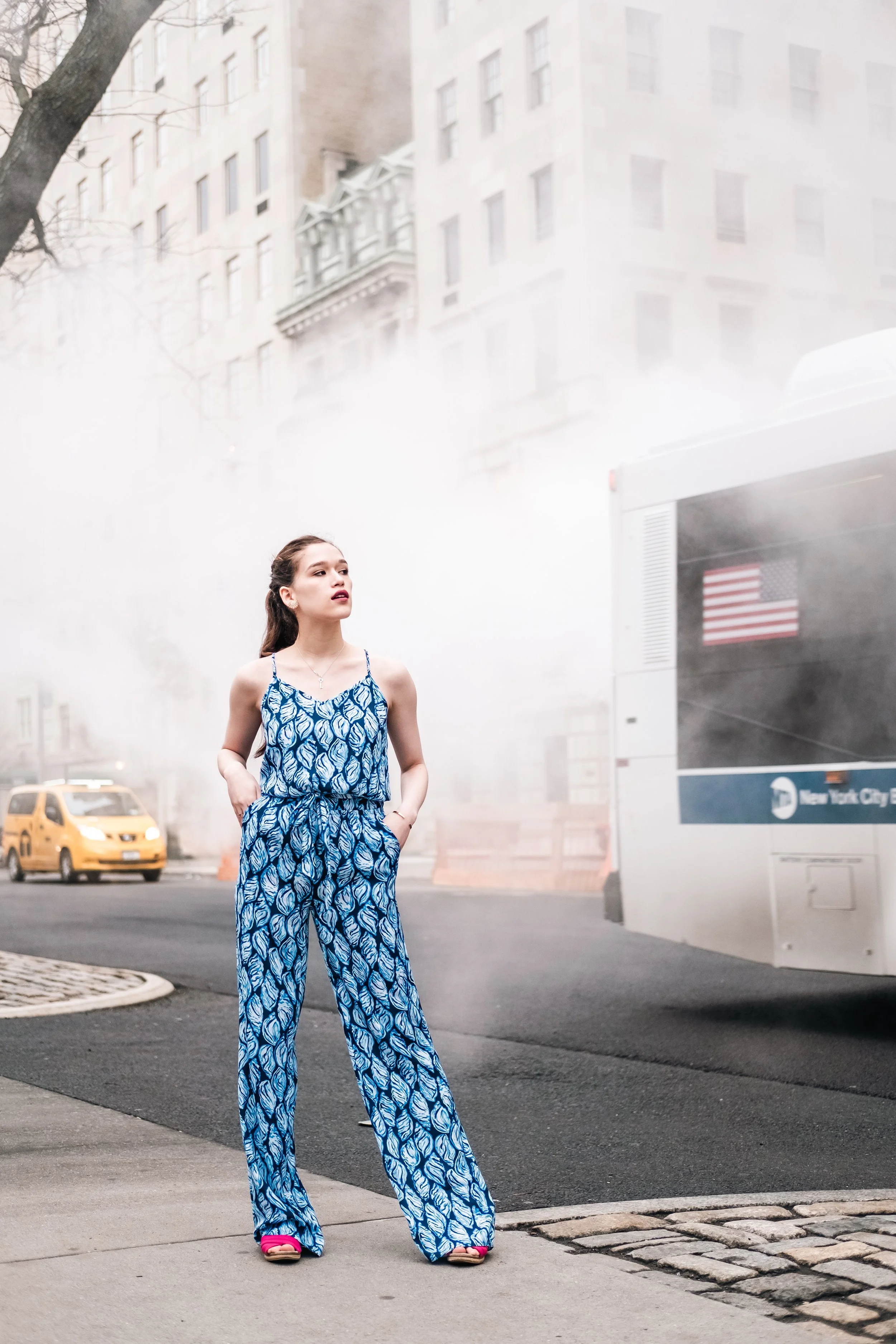 A woman wearing a matching blue and white patterned sleeveless jumpsuit and pink high-heeled shoes stands on a city sidewalk. She has her hands in her pockets and is looking to her left. Smoke or fog is coming from a building behind her, with an Amer