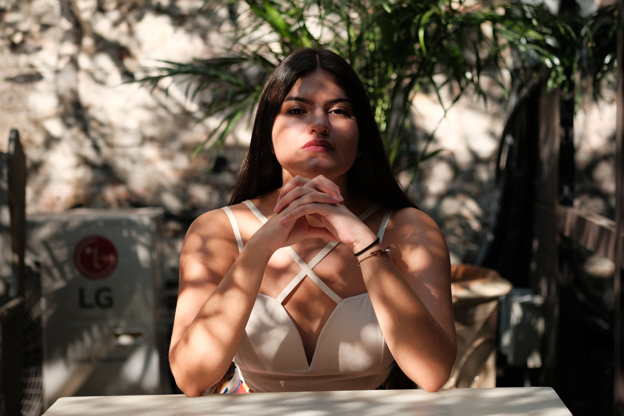 A young woman with long dark hair and light skin sitting outdoors at a table, with her hands clasped under her chin, looking directly at the camera. She is wearing a white strappy top, and sunlight is casting shadows on her face. The background featu