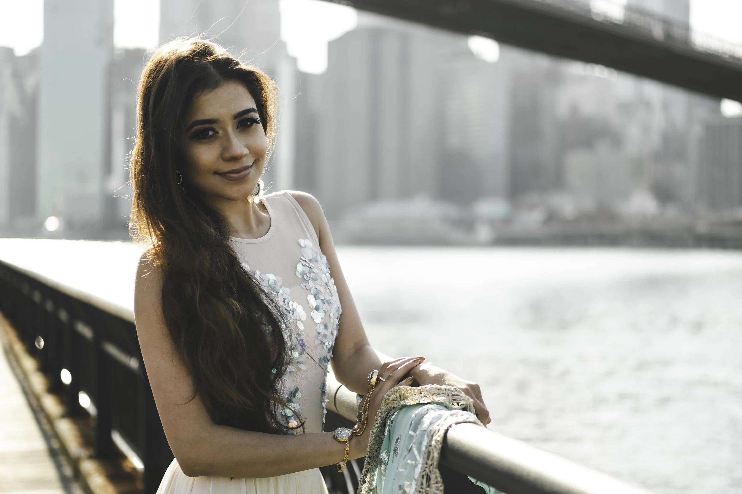 A woman with long dark hair, wearing a sleeveless top with sequins, sitting on a bridge with a city skyline and water in the background.
