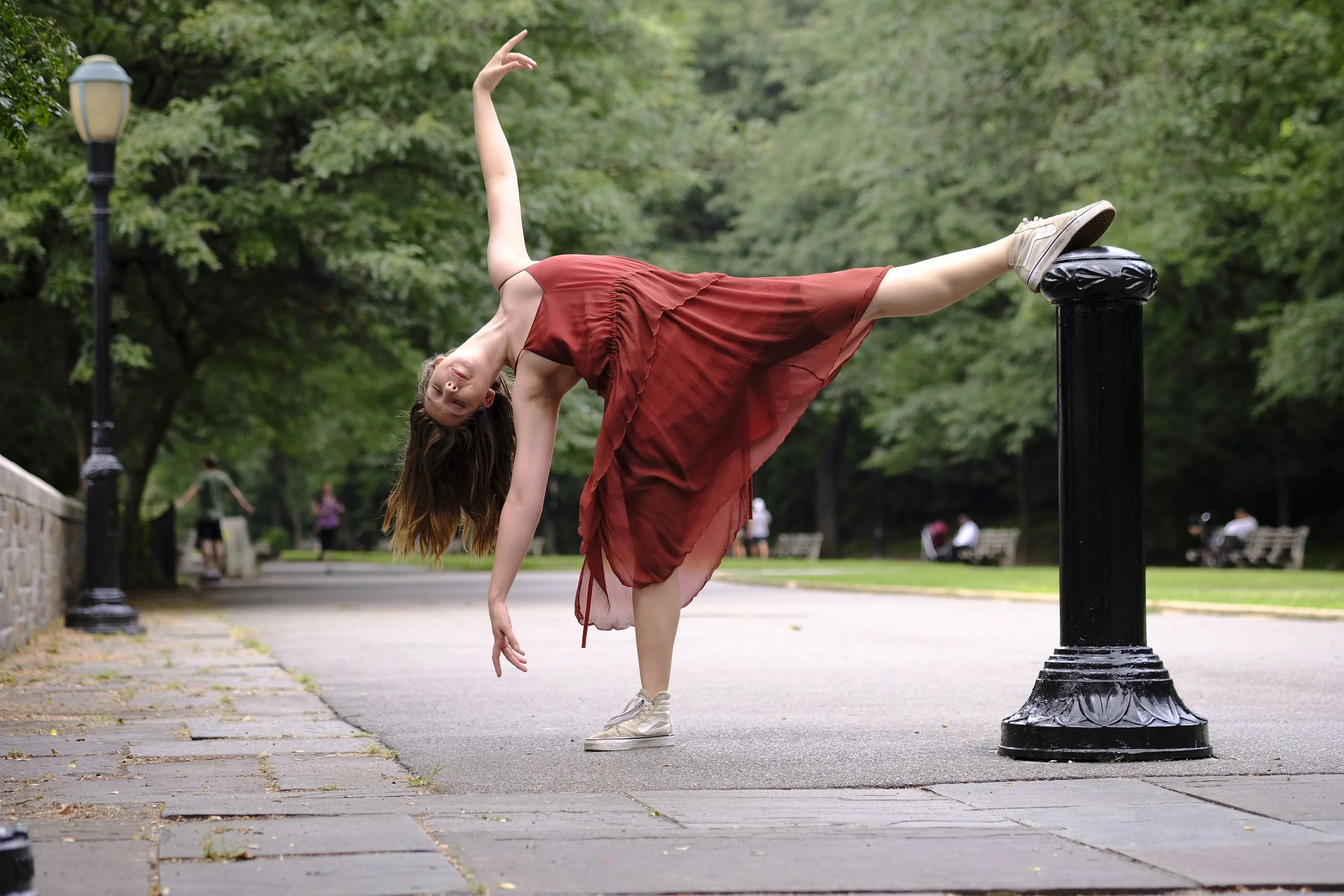 A woman in a red dress balances on her left foot with her right foot resting on a black street lamp post in a park. She leans forward with her left hand touching the ground and her right arm raised above her head. Green trees and park benches are vis