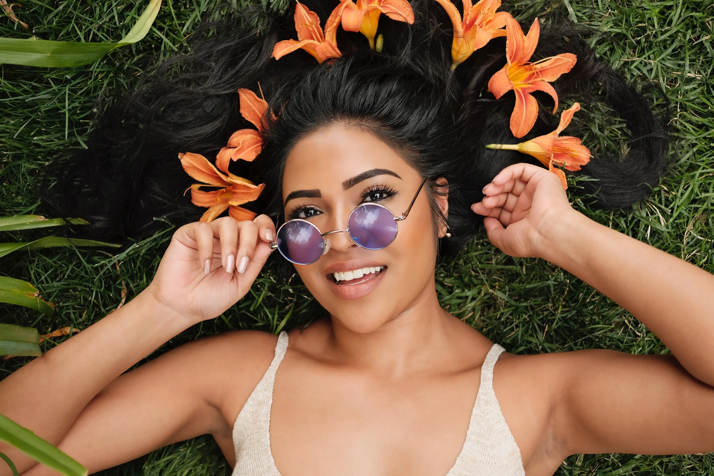 A woman with long black hair lying on grass, surrounded by orange lilies, holding sunglasses, smiling, wearing a white tank top.