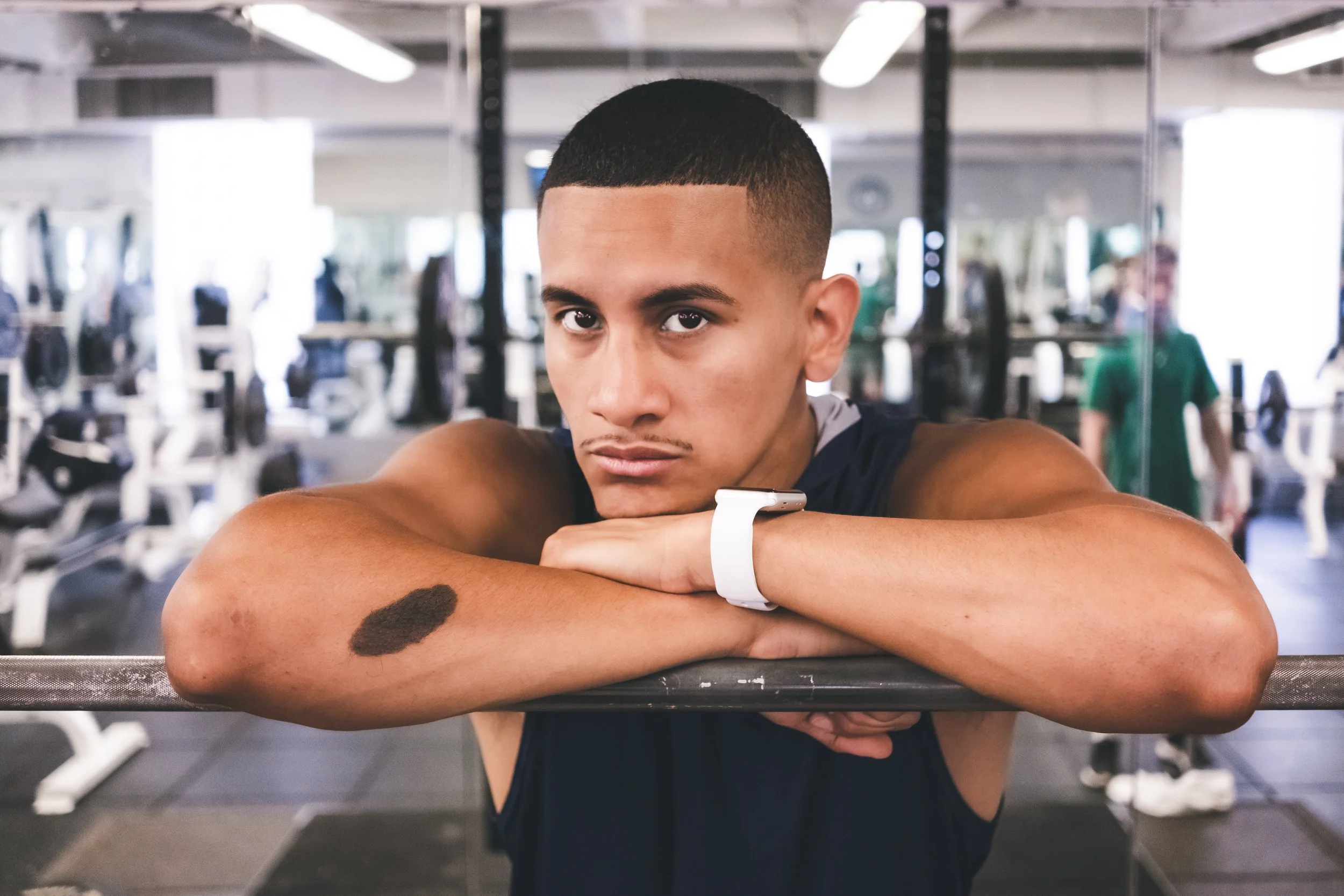 A young man with short hair and a tattoo on his arm taking a selfie at the gym, resting his arms on a flat surface with gym equipment in the background.