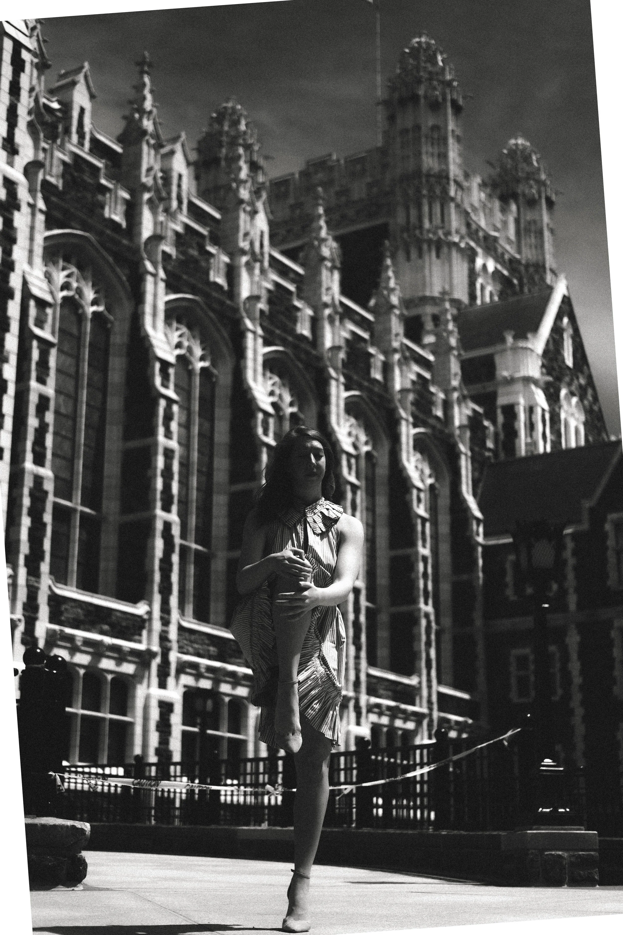 A woman in a dance pose standing on one leg with her arms crossed in front of her chest, in front of a large ornate gothic-style building.