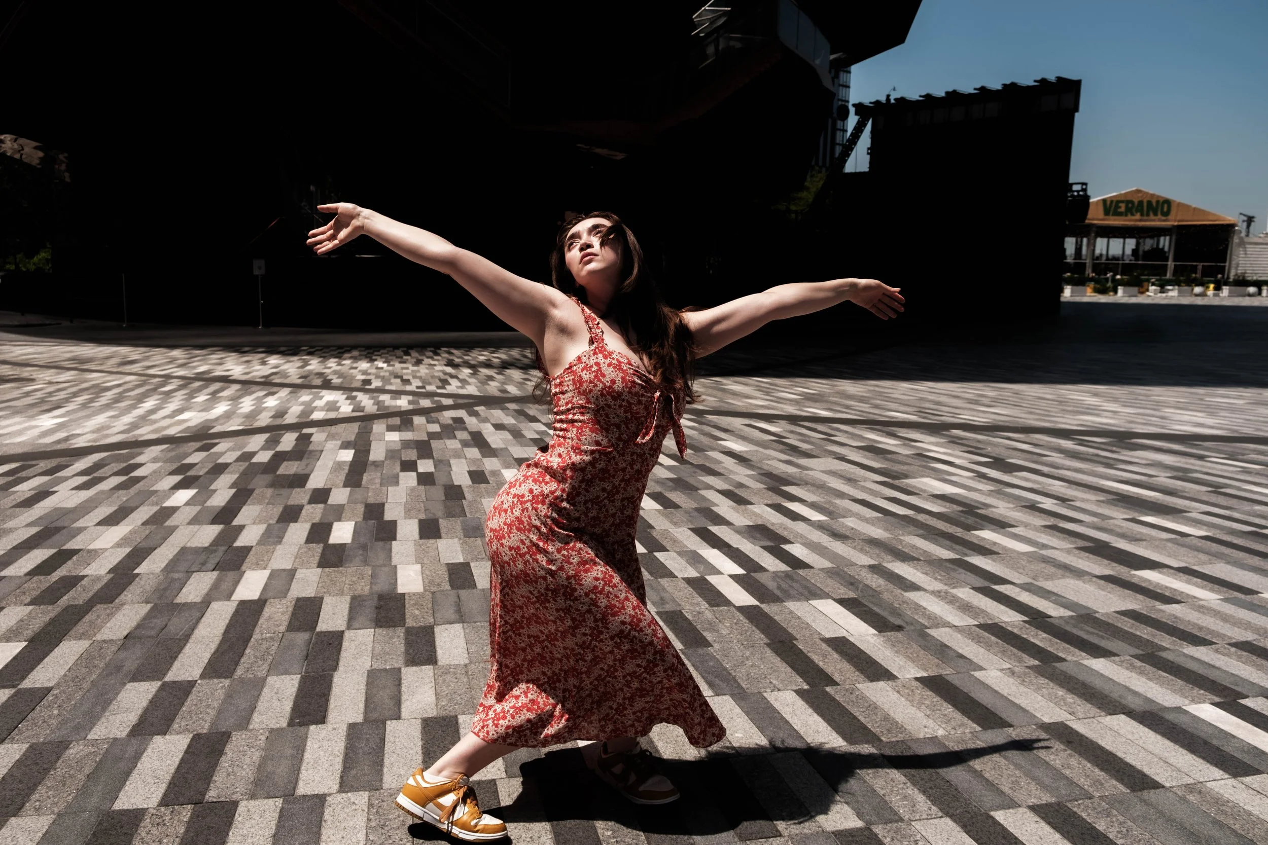 A woman in a red floral dress poses outdoors in front of a dark structure and cityscape, extending her arms wide and looking upward.