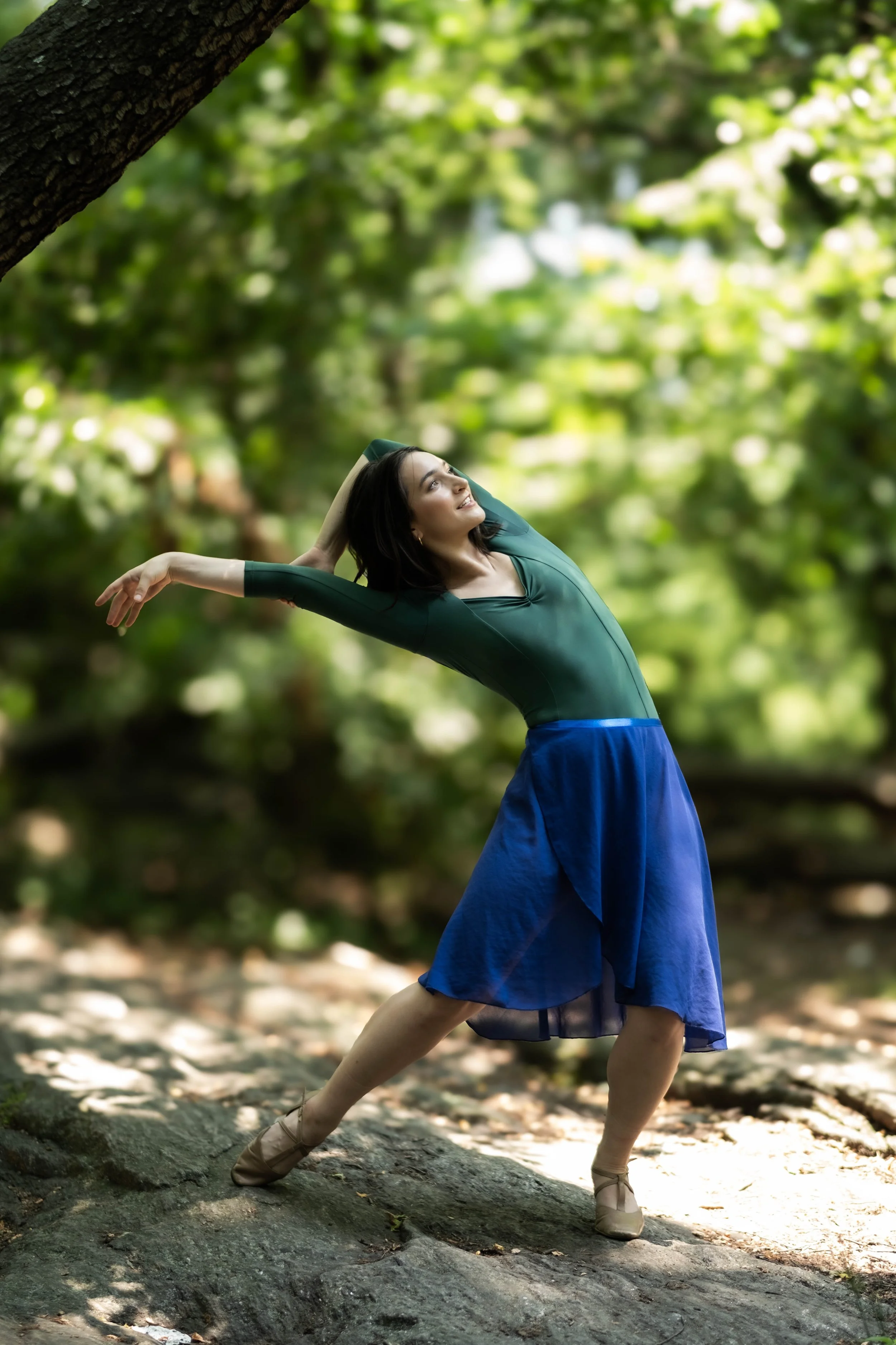 A woman in a green dance dress and ballet shoes practicing ballet outdoors on a rocky surface with trees and greenery in the background.