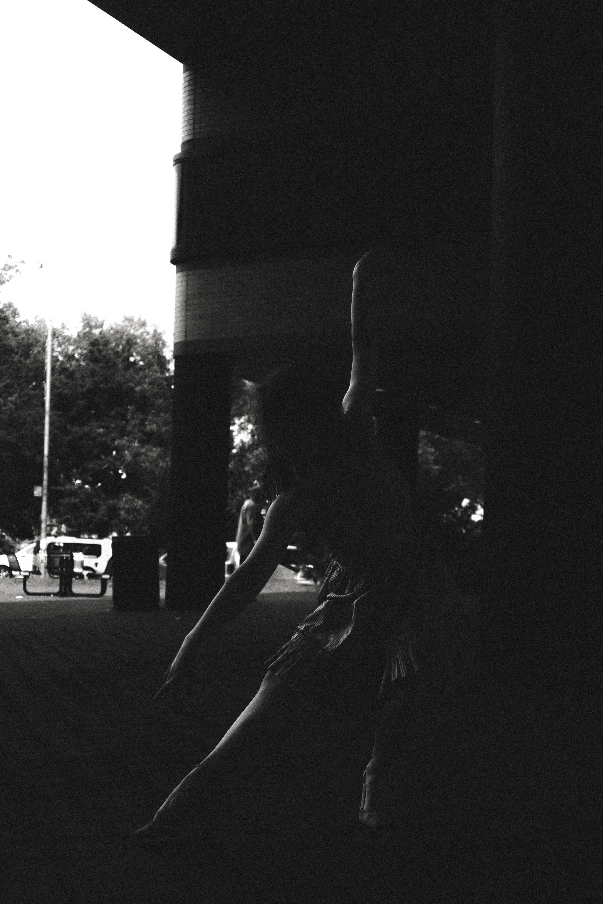 Black and white photo of a person skateboarding under a bridge or overpass, with trees and parked cars in the background.