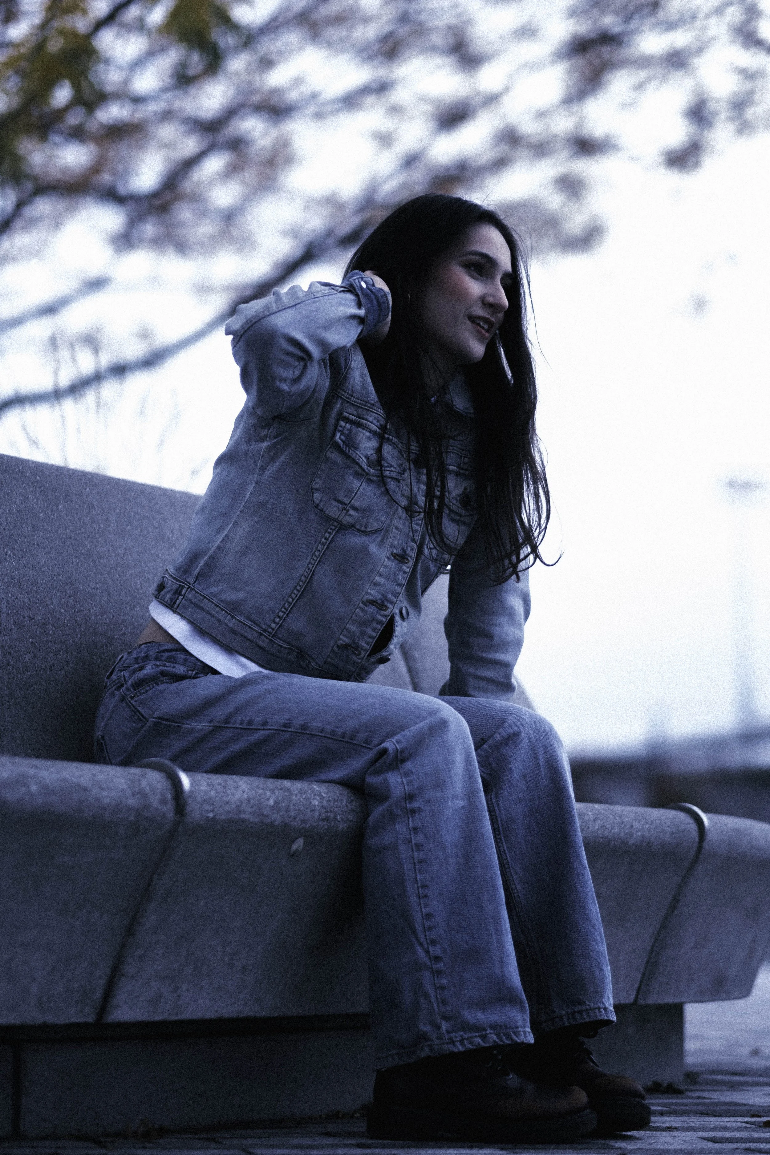 A young woman with long dark hair wearing a denim jacket and jeans sitting on a concrete bench outdoors, adjusting her hair or ear, with trees and sky in the background during dusk.