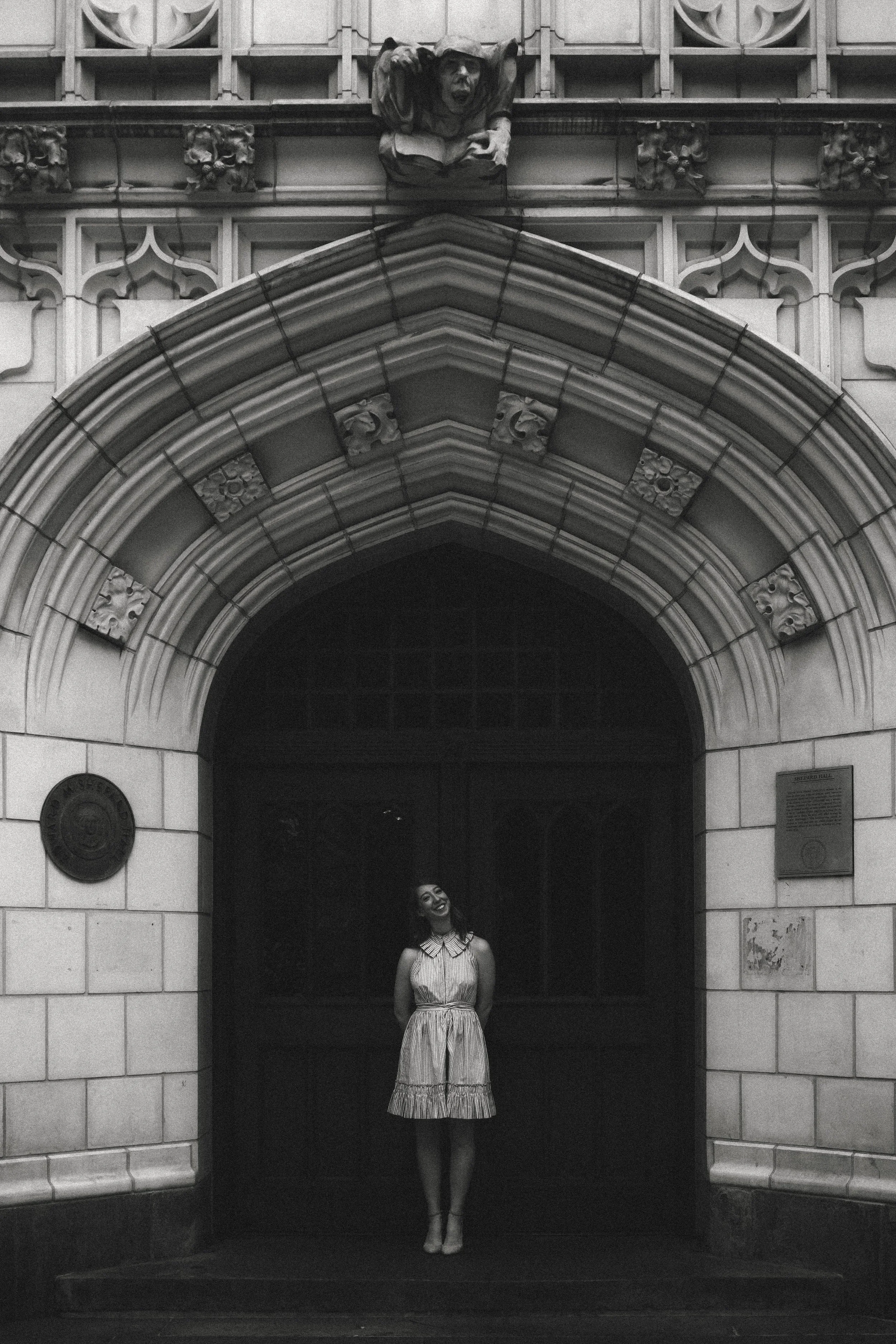 Black and white photo of a woman in a striped dress standing in front of a dark archway, smiling. Above the archway, there is a sculpted figure of a person with an open mouth holding a book, and decorative architectural details.