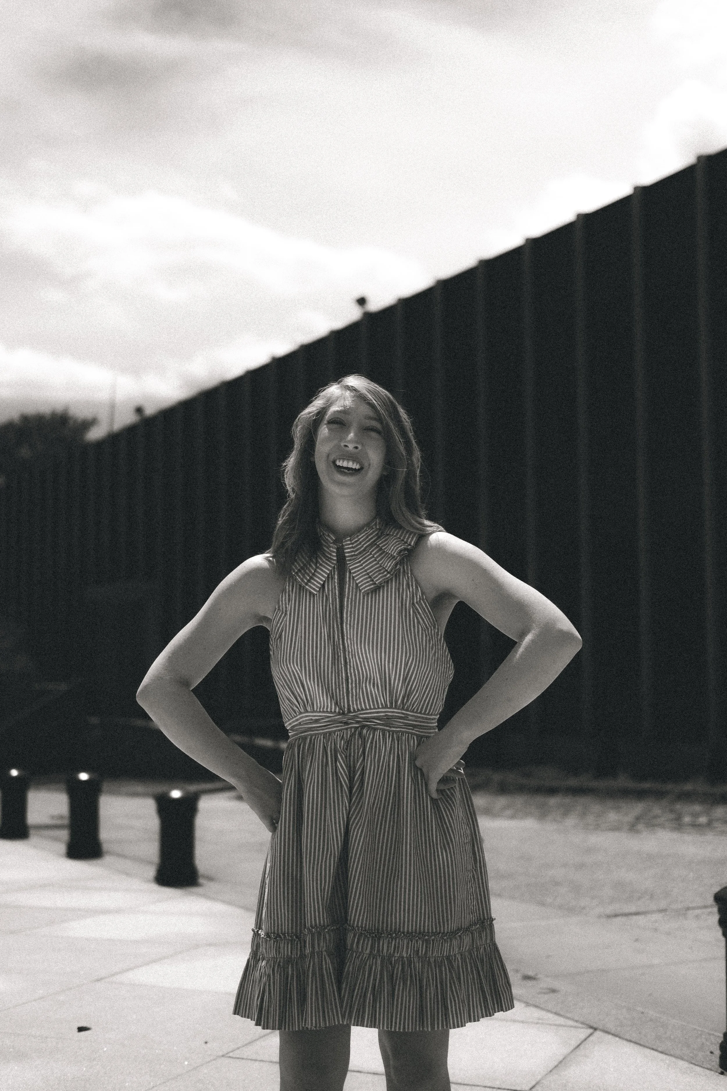 A young woman standing outdoors in front of a black fence with her hands on her hips, smiling and looking up. She is wearing a sleeveless, striped dress with a ruffled hem.