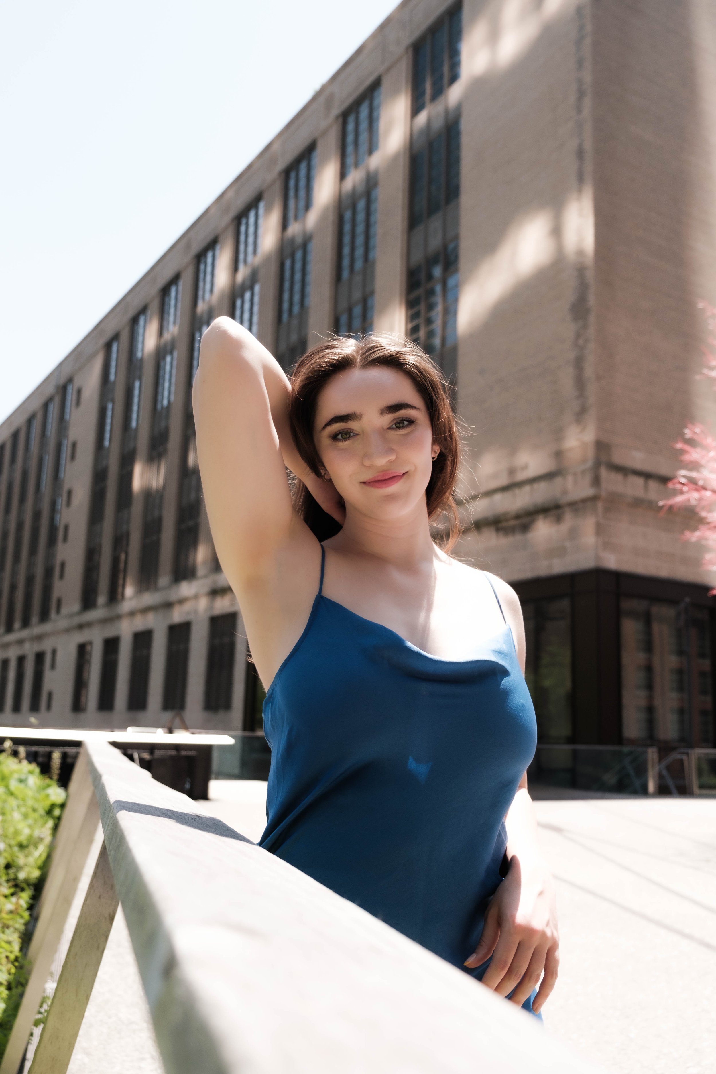 Young woman with short brown hair wearing a blue dress, smiling with one arm raised behind her head, standing outdoors near a white bench with a large building in the background.