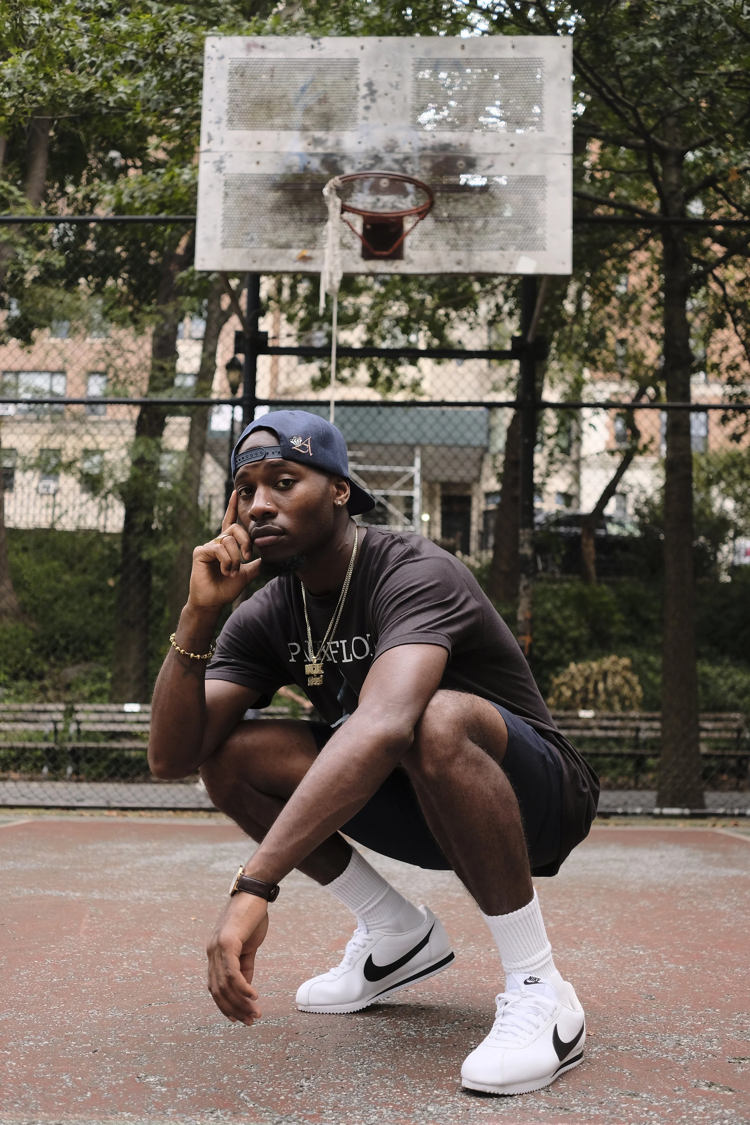 A young man crouching on a sports court, dressed in a black t-shirt, black shorts, white socks, and white Nike sneakers, with a backwards cap, sitting in front of a basketball hoop on an outdoor court.