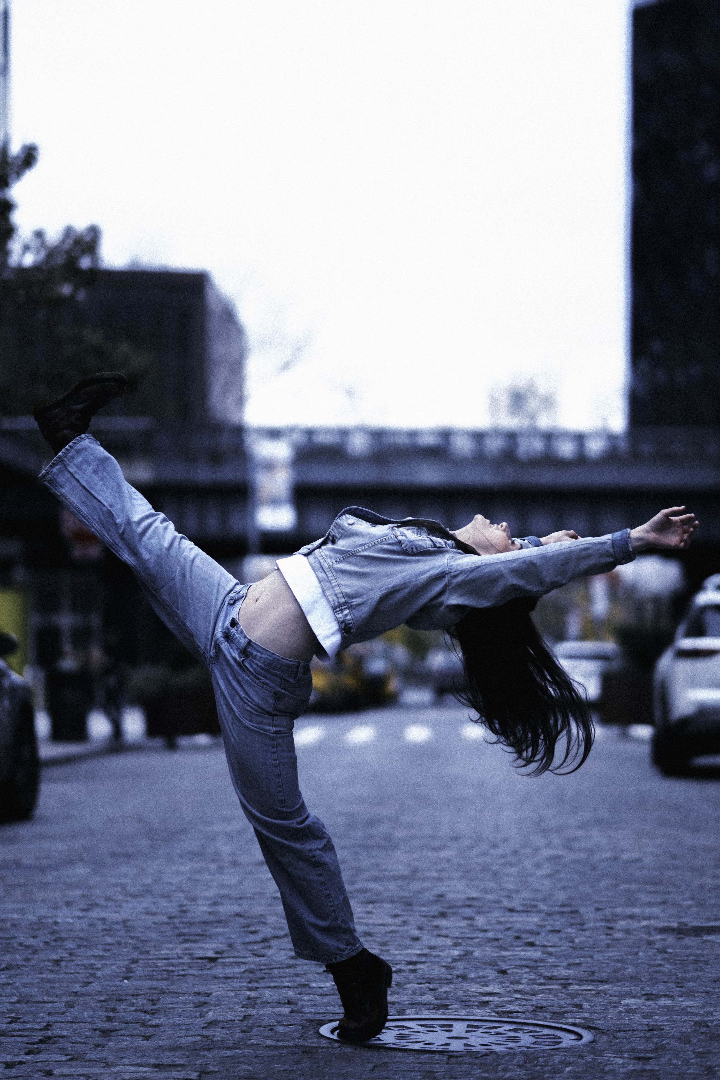 A woman in a denim jacket and jeans performing a balancing yoga pose on a city street during evening, with her arms stretched forward and one leg raised behind her, standing on a manhole cover.