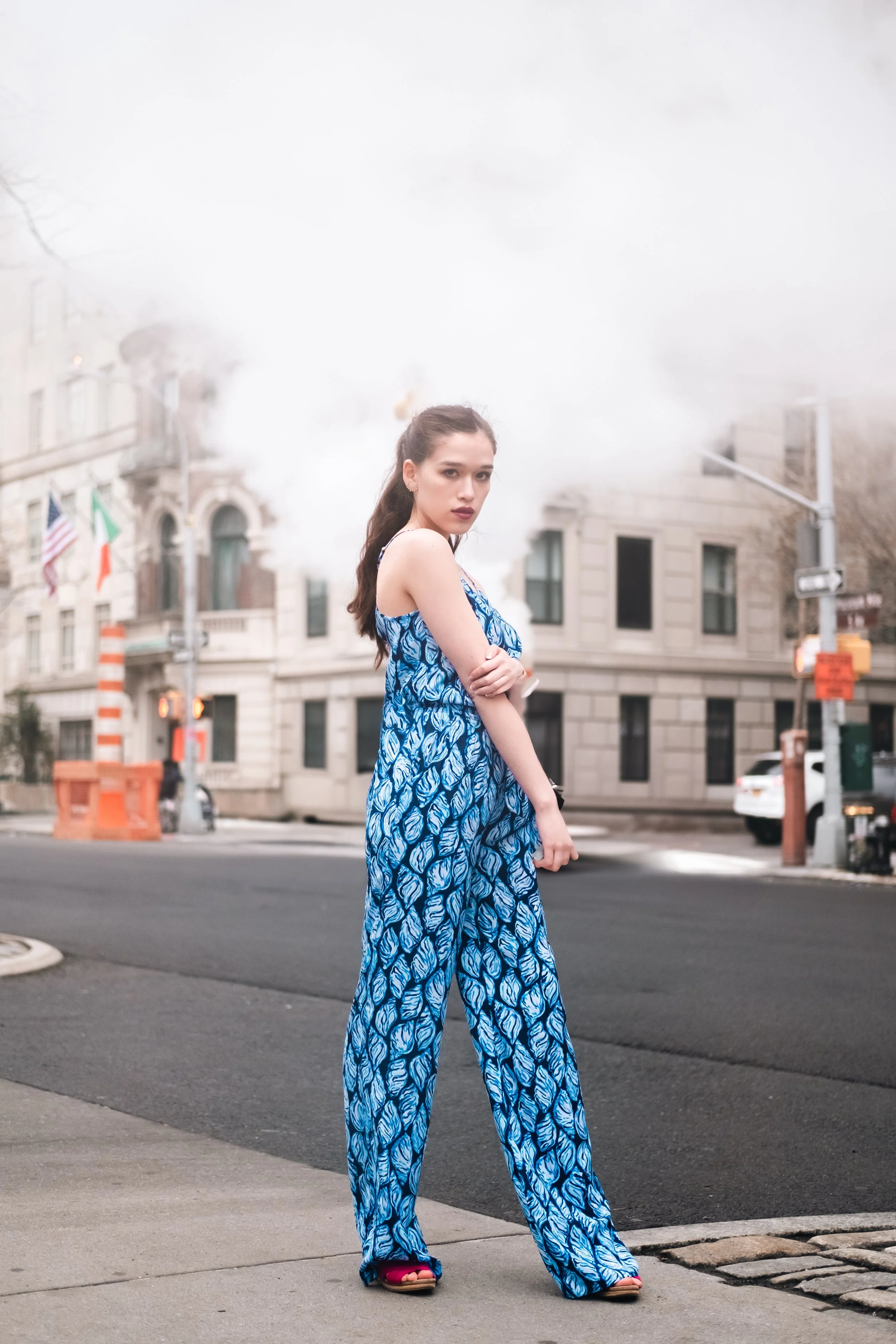 A young woman with long dark hair, wearing a blue patterned jumpsuit and pink shoes, standing on a city street corner with buildings and traffic cones in the background.