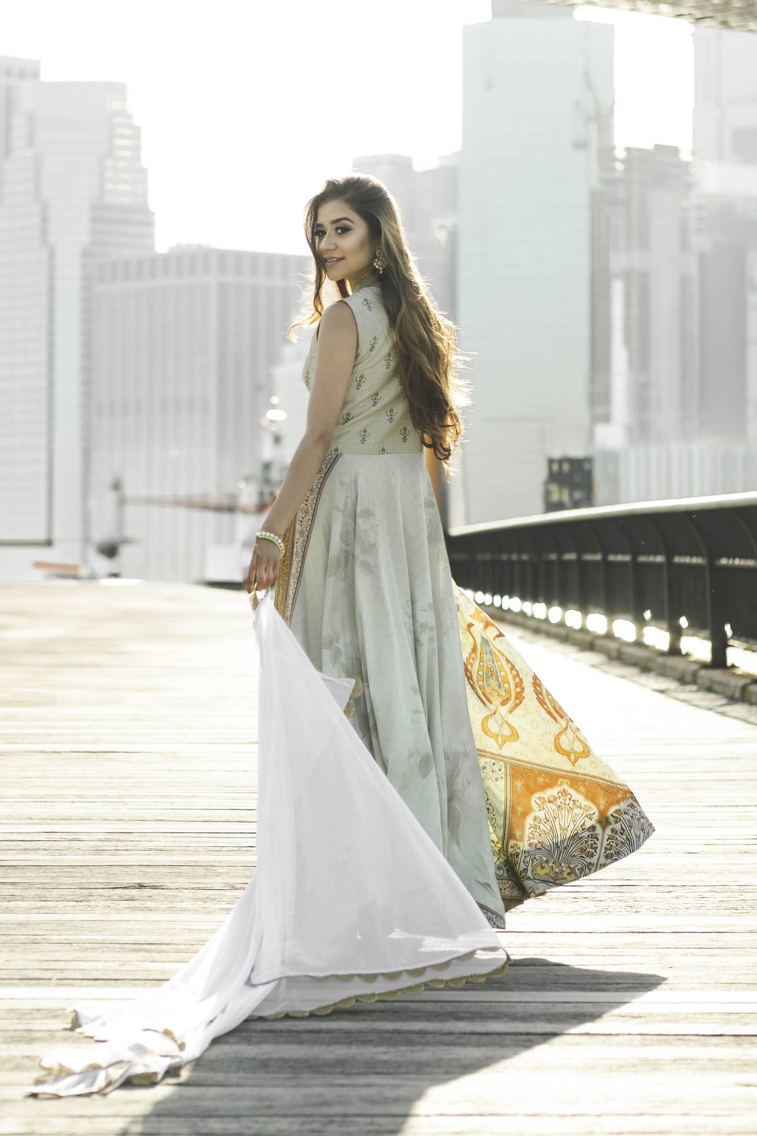 A woman in traditional Indian attire standing on a wooden deck in an urban area during daylight, with tall modern buildings in the background.