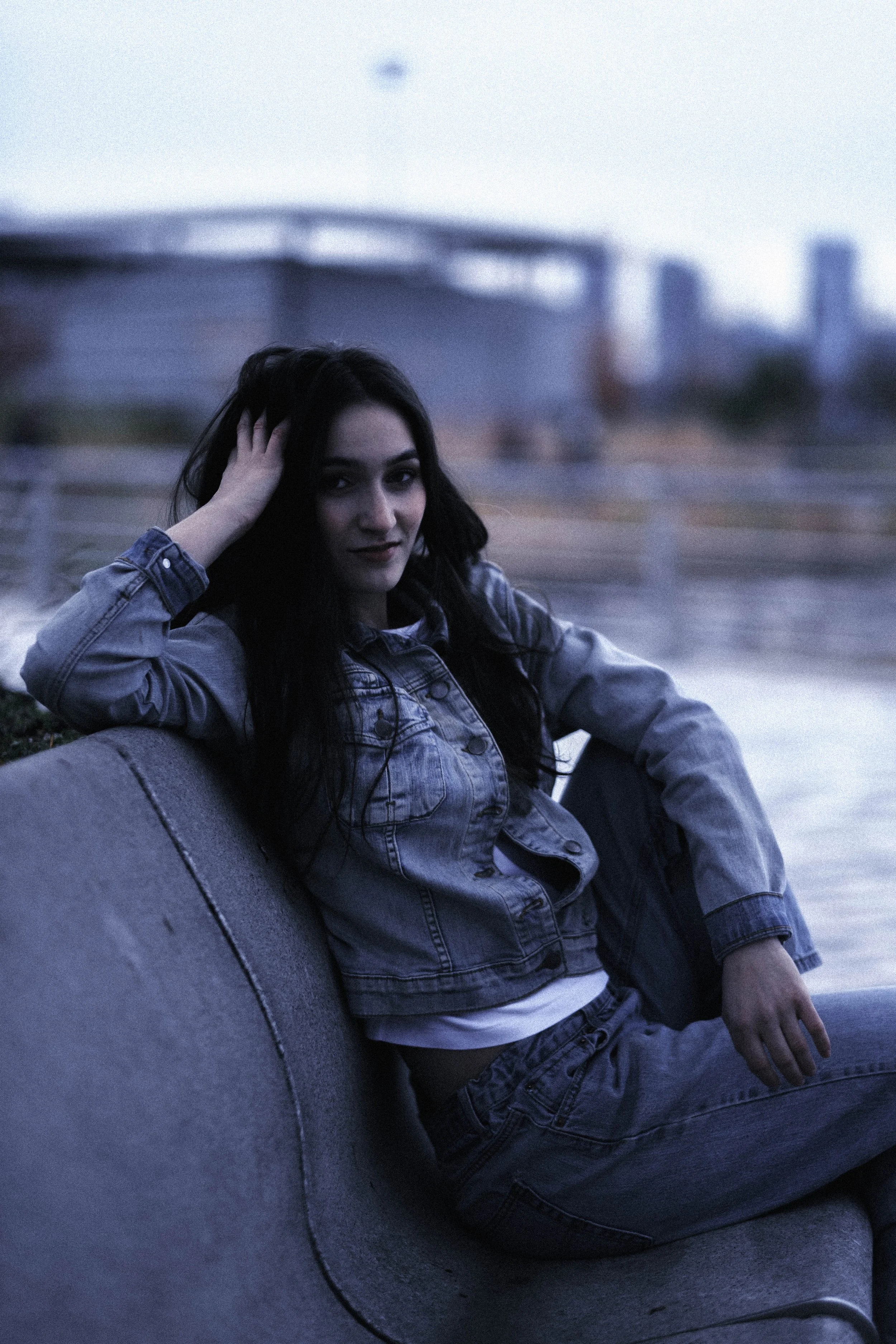 A young woman with long dark hair wearing a denim jacket and shorts, sitting on a curved city bench at dusk with a blurred cityscape in the background.