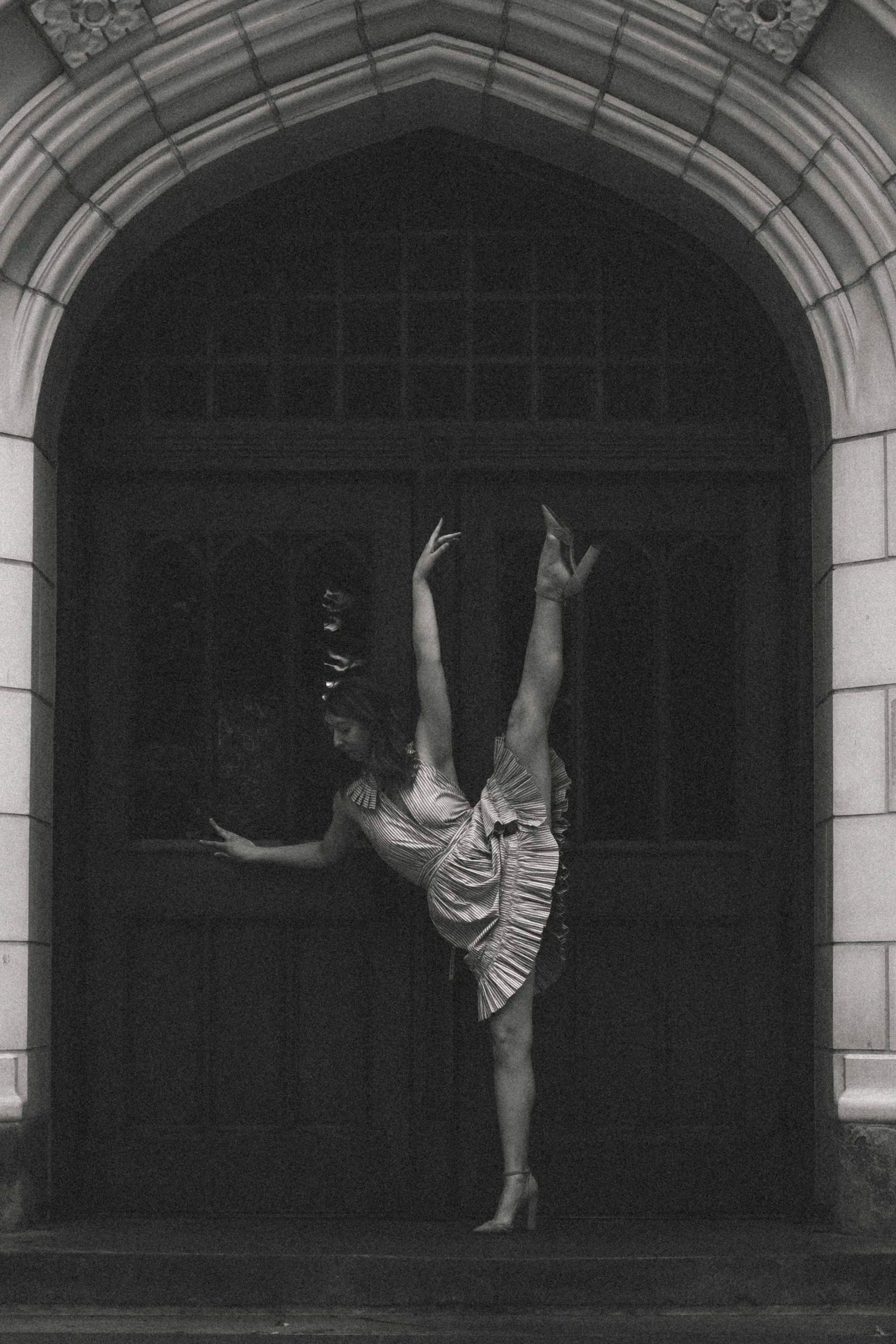 A dancing woman in a striped dress performs a ballet pose on one leg in front of a dark arched doorway, with hands raised and head bowed.