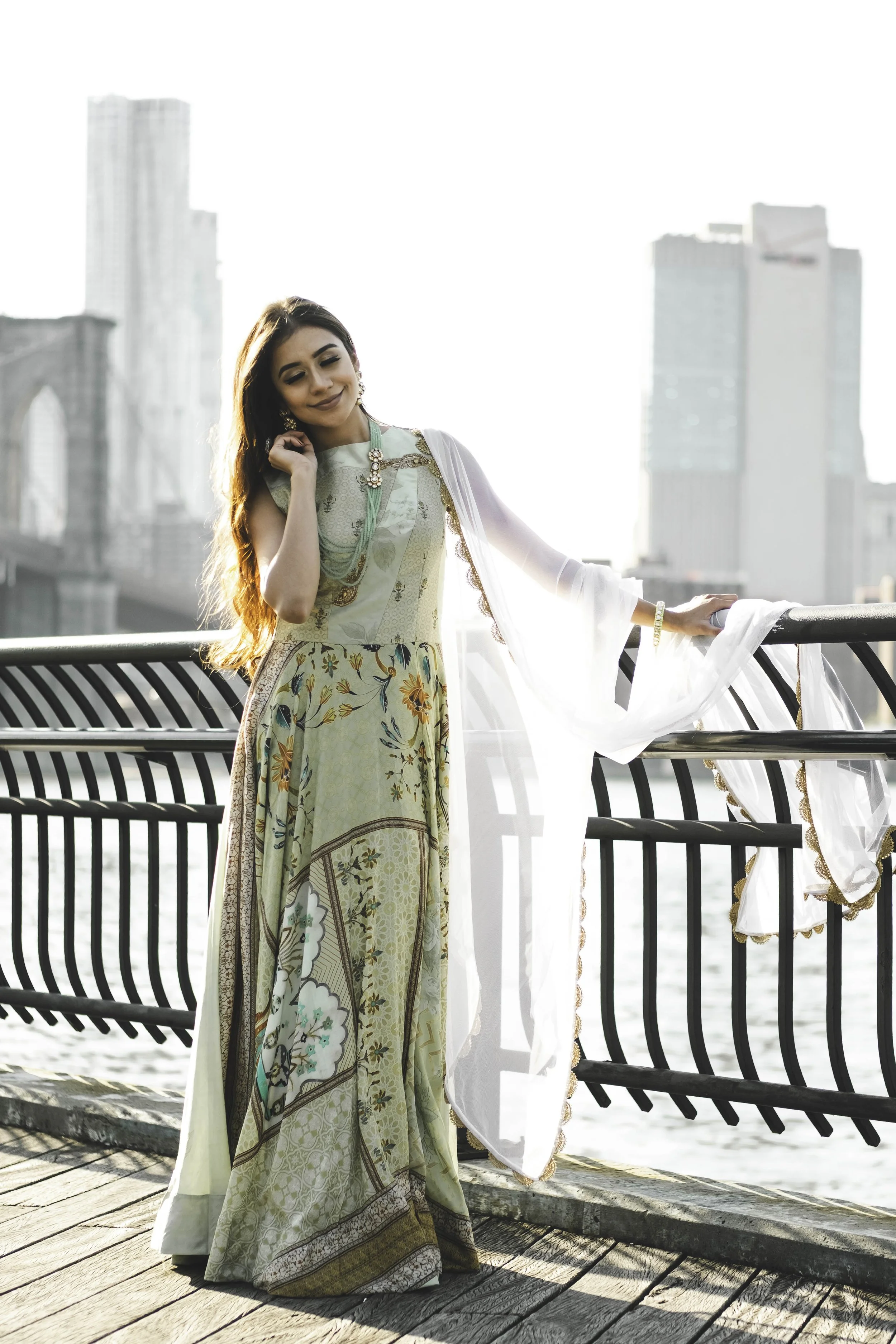A woman dressed in a traditional, colorful, floral and patterned Indian dress stands on a wooden boardwalk by the water, with a city skyline and skyscrapers in the background.