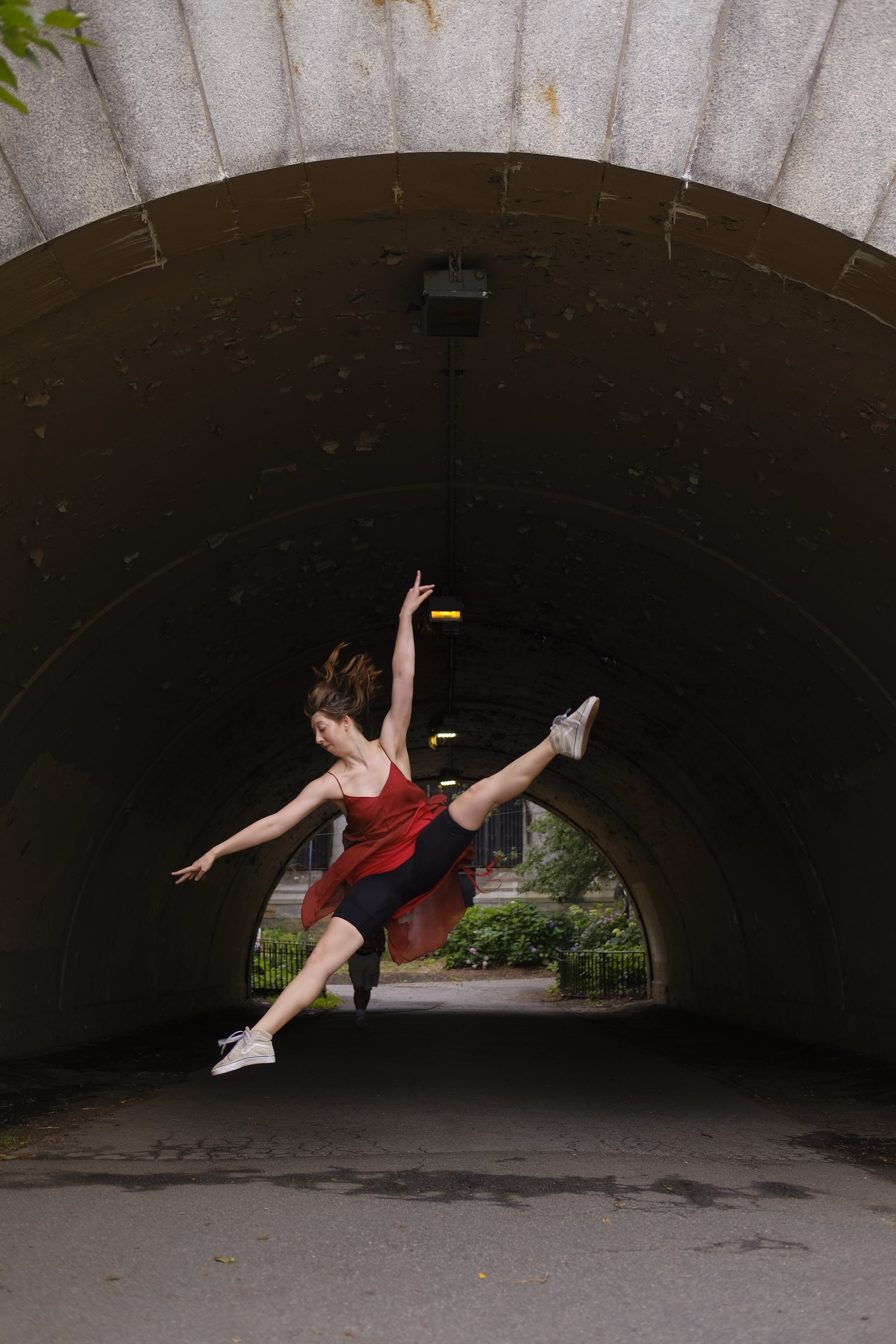A dancer in a red dress performing a leap in a tunnel with a dark ceiling and bright outside greenery.