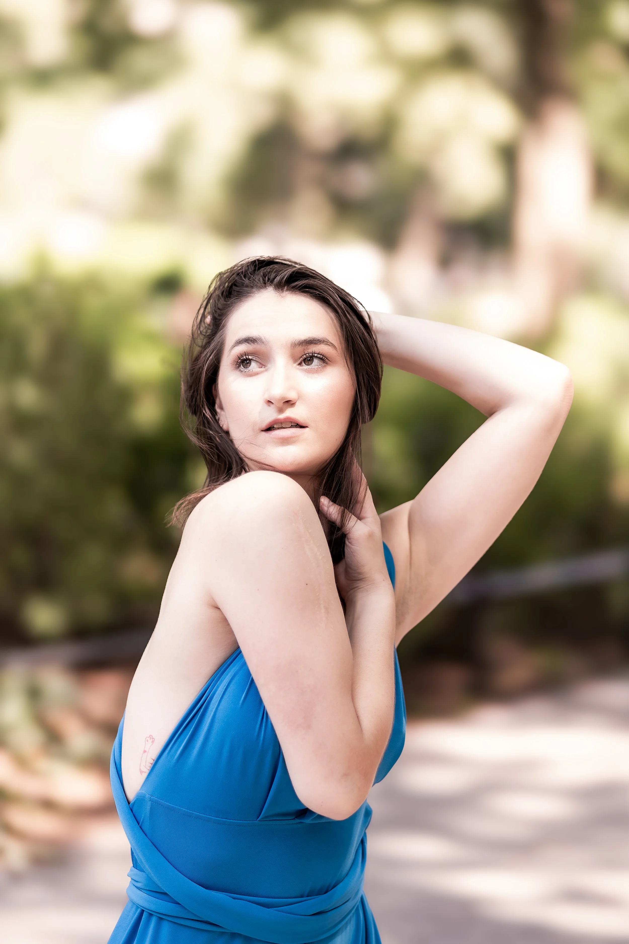 A young woman with brown hair in a blue dress posing outdoors with blurred green foliage background.