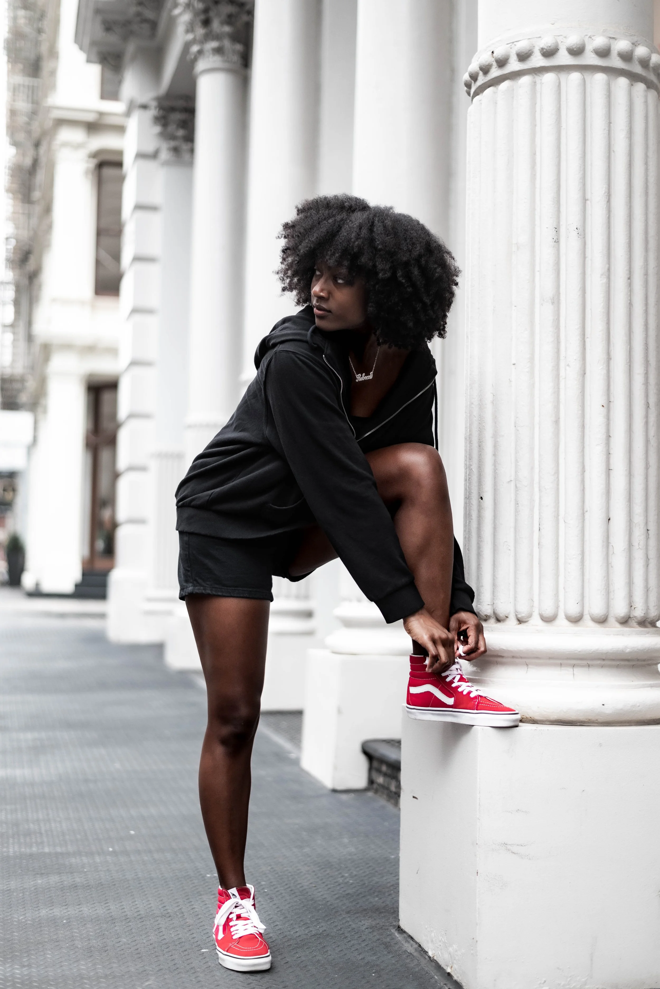 A young woman with afro hair is stretching outside a building with tall white columns, wearing a black hoodie, black shorts, and red and white sneakers.