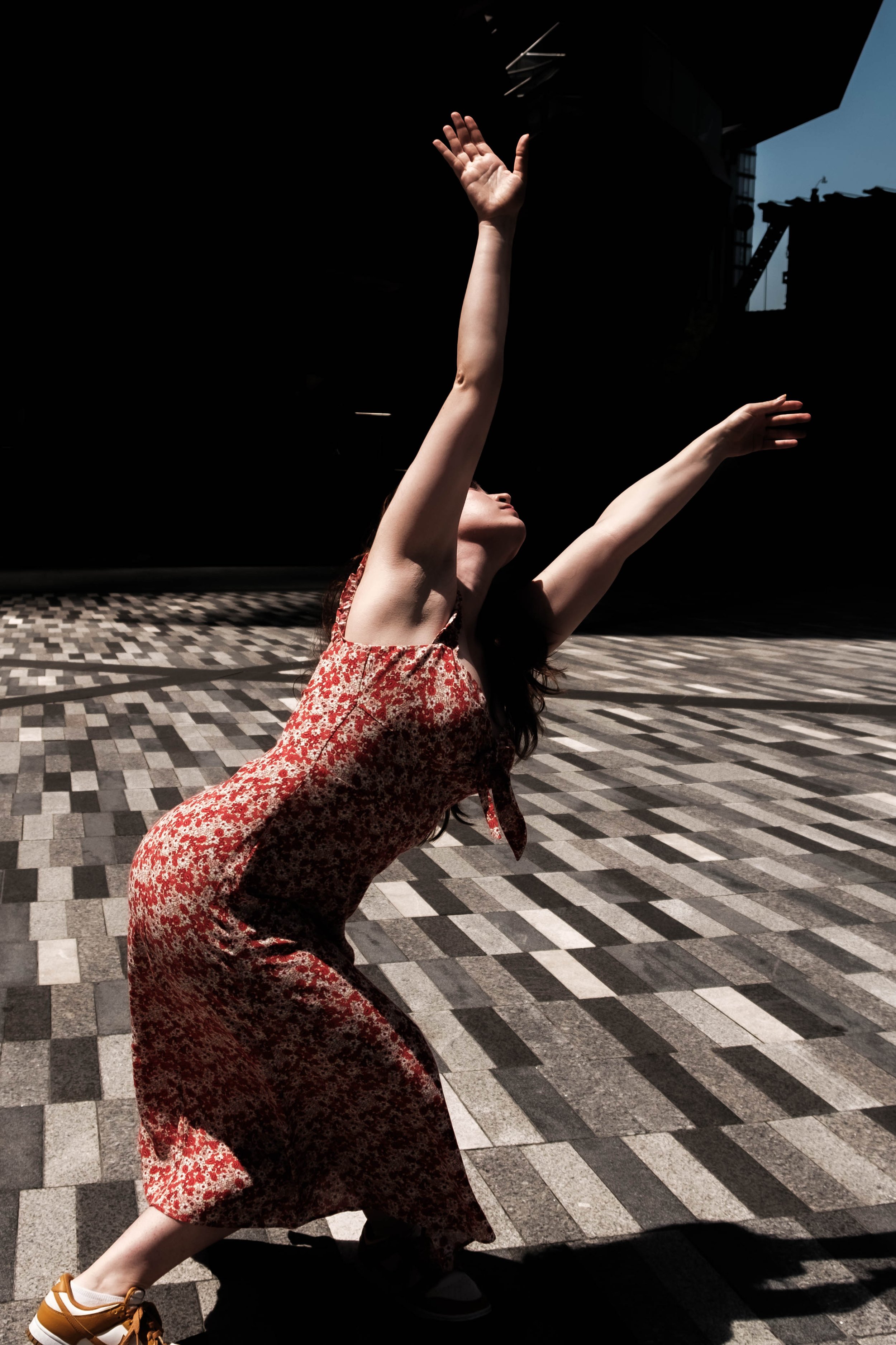 A woman in a red floral dress bending backward on a geometric patterned pavement, with arms raised and head tilted back, outdoors with dark background and blue sky.