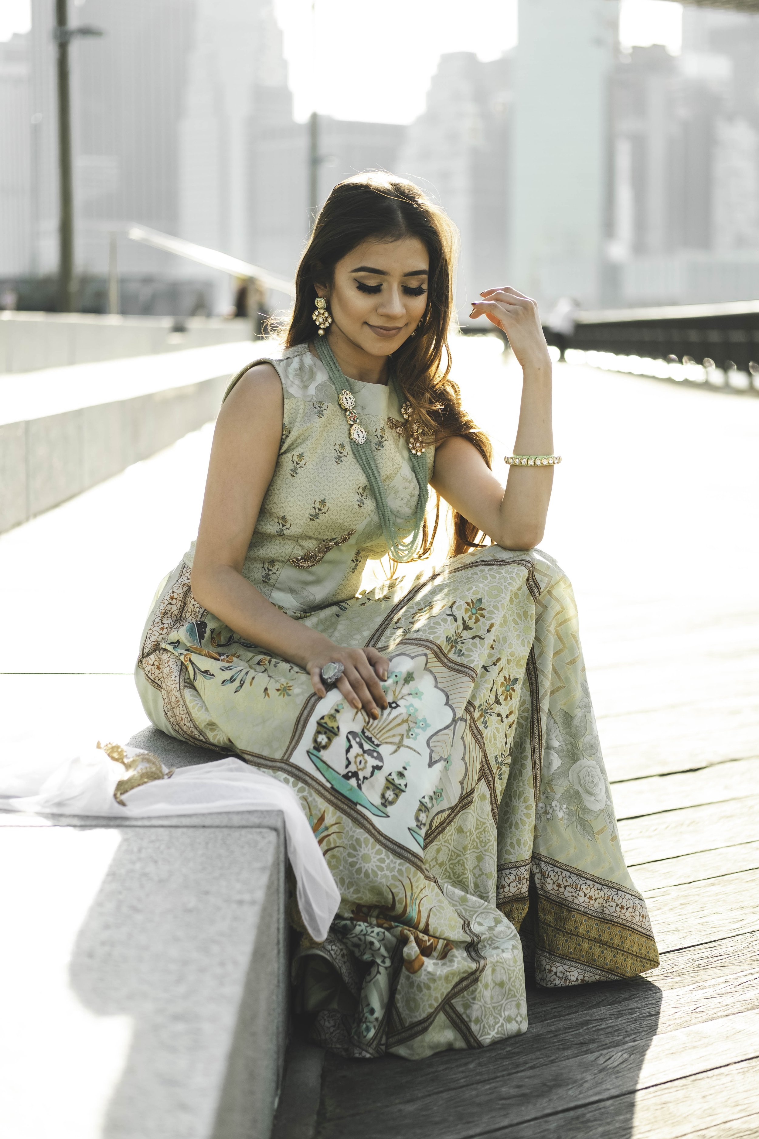 A woman dressed in traditional attire sitting on a wooden dock near a city skyline. She has long hair, wears earrings and a bracelet, and is holding a decorative item in her lap. The setting is sunny with a bright background.