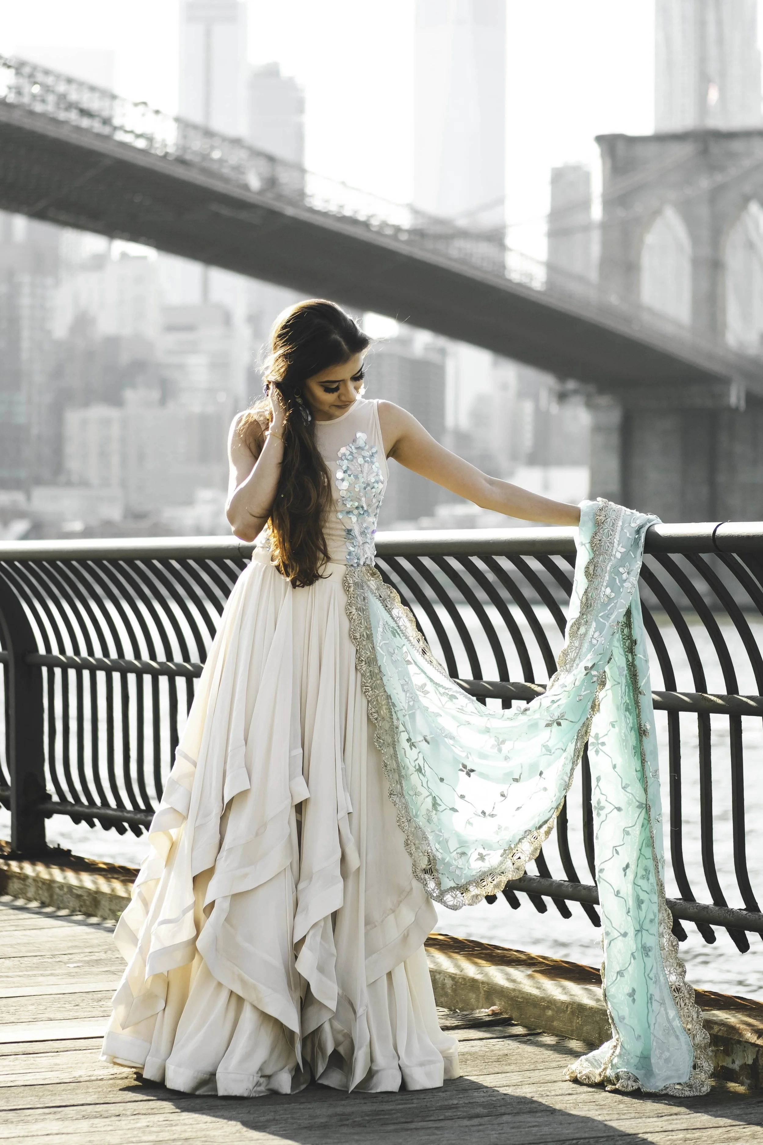A woman in a white gown with embroidery, holding a turquoise embroidered fabric, standing on a bridge with a cityscape and a bridge in the background during daylight.