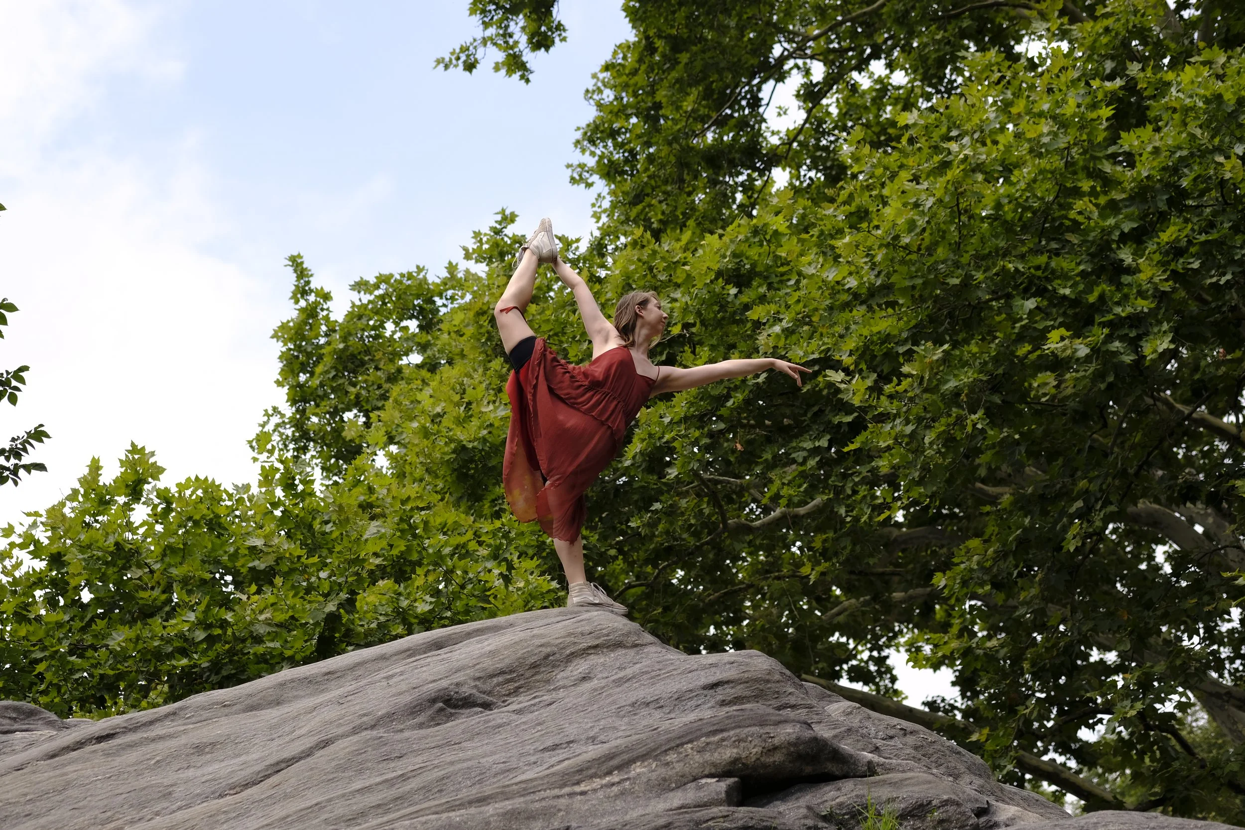 A woman in a red dress and white sneakers practicing a one-legged balancing pose on a rock in a park with green trees around.