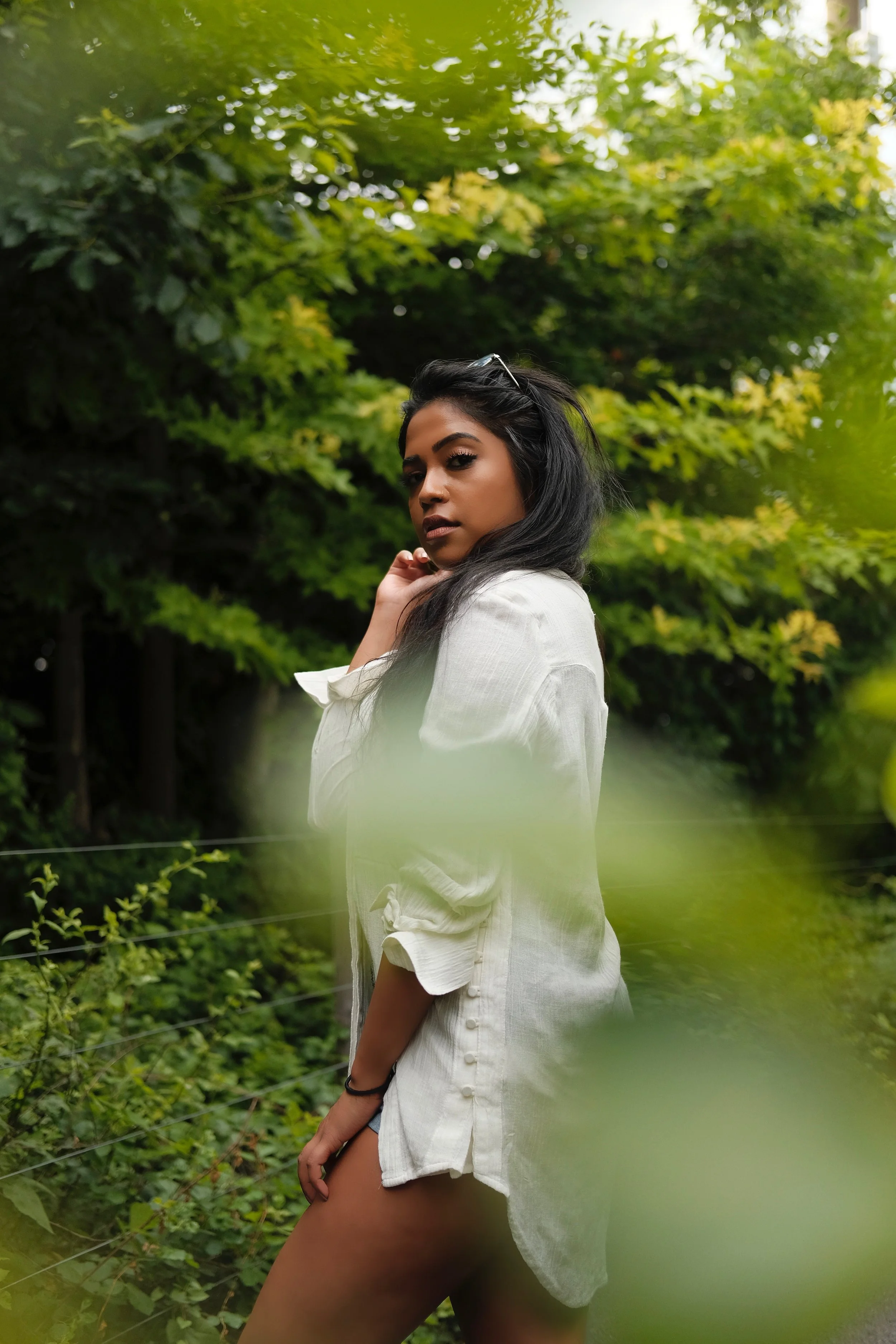 A woman standing outdoors in a natural setting with green foliage in the background. She is wearing a white shirt and has dark hair, with a contemplative expression on her face.