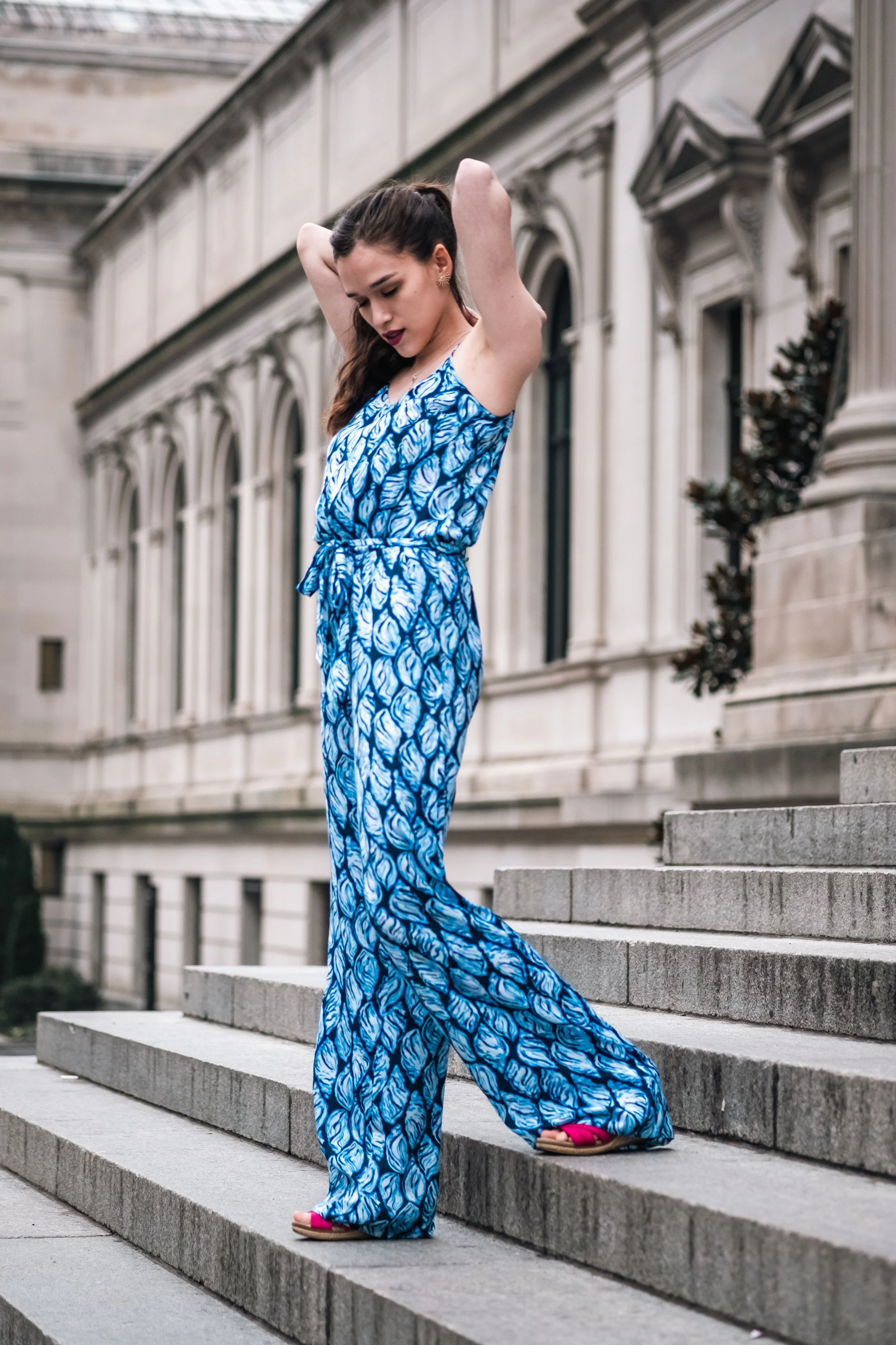 A young woman in a blue patterned jumpsuit standing on outdoor concrete stairs, adjusting her hair, with dark hair and wearing pink sandals, in front of a historic building.