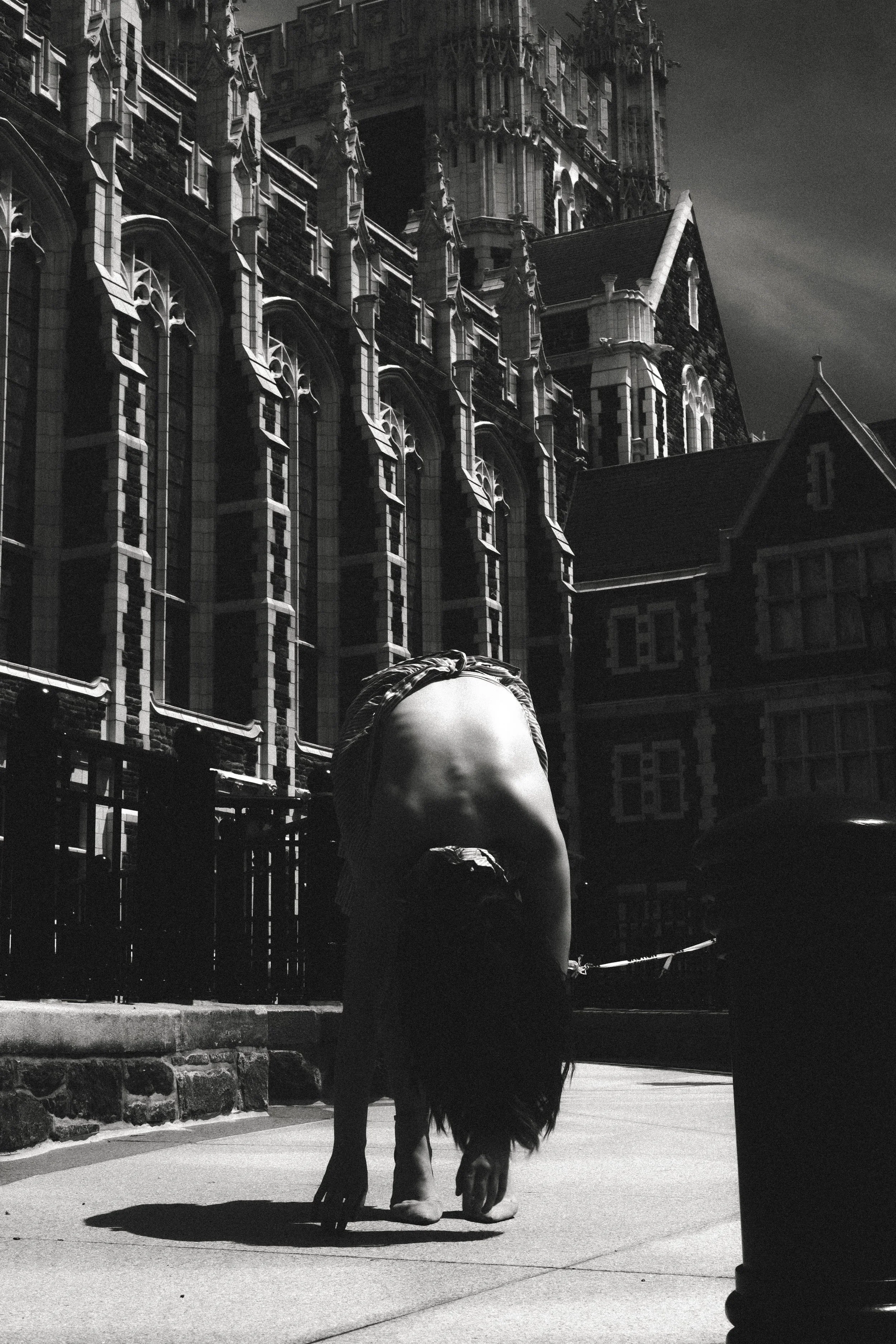 A person with long hair walks a horse on a city sidewalk, with a large gothic-style building in the background, black and white photo.