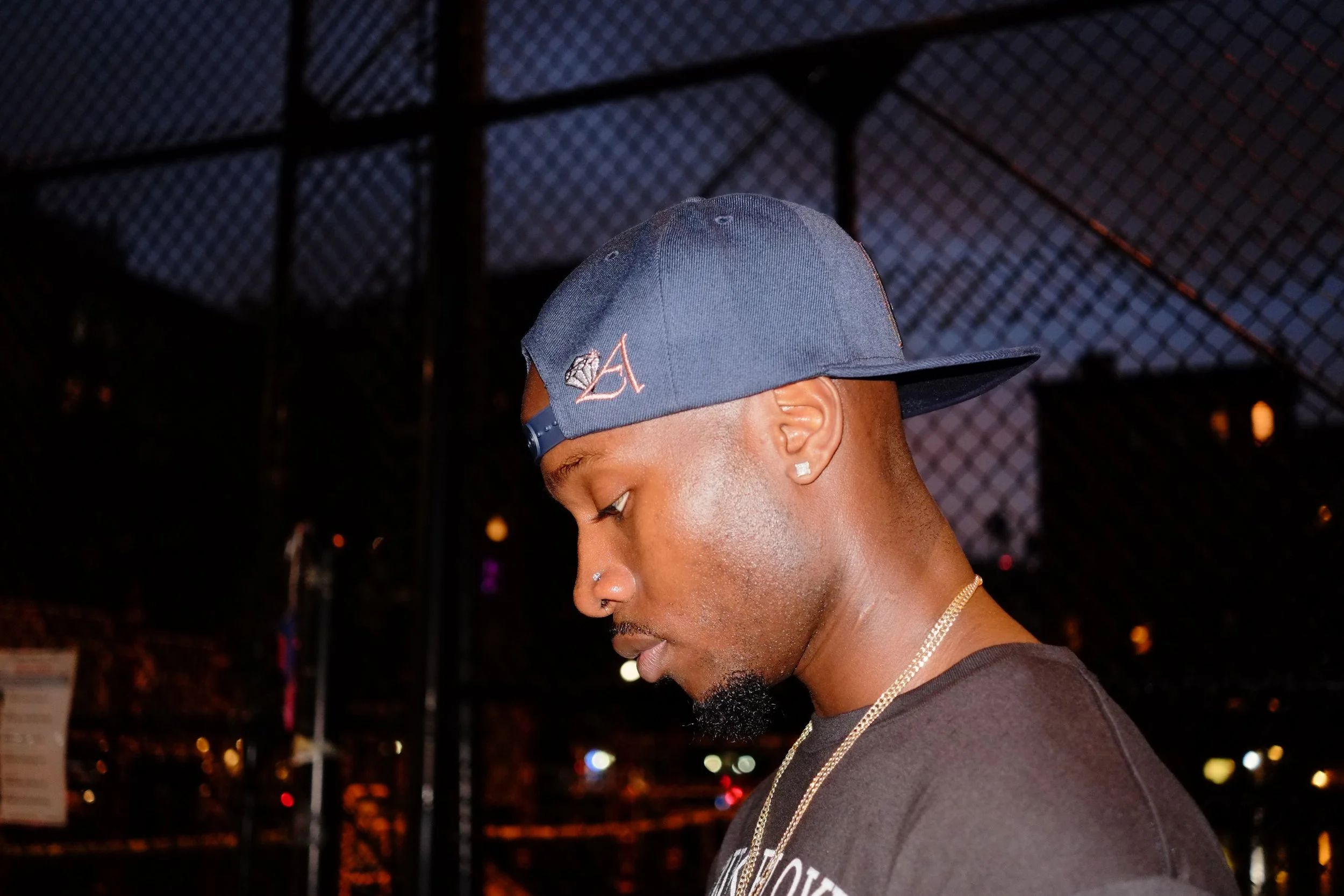 Close-up of a man with a cap and earrings, looking down, at night with a blurred outdoor background.