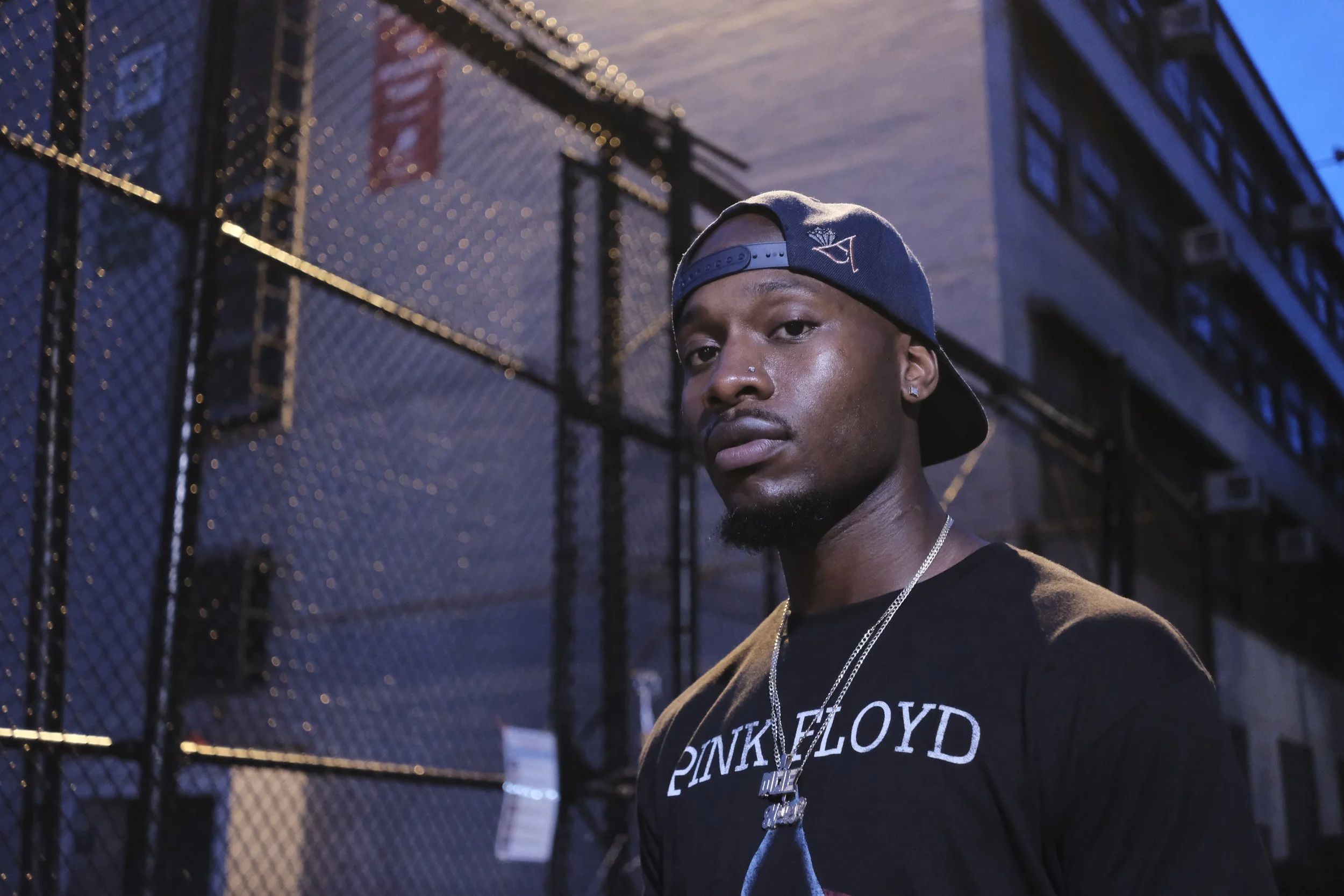 Close-up of a young man wearing a black Pink Floyd t-shirt, a silver chain, and a navy blue baseball cap backward, standing in front of a chain-link fence at dusk.