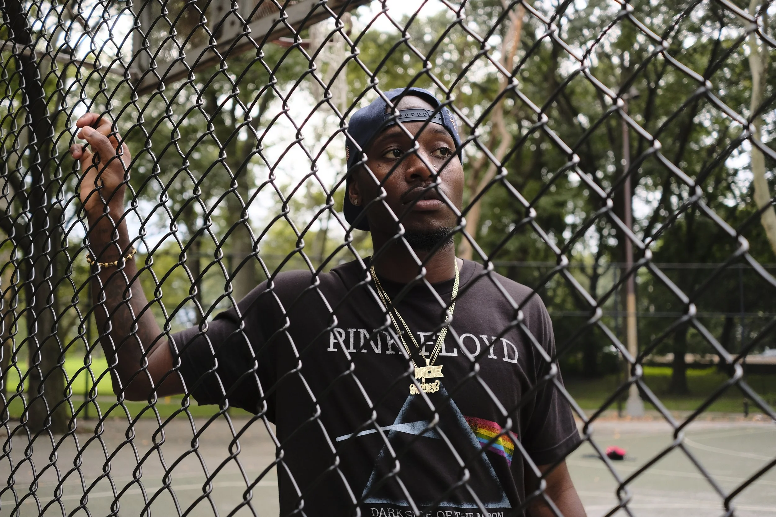 A man wearing a black "RIP Floyd" t-shirt, multiple chains, and a black baseball cap backwards stands behind a chain-link fence on a tennis court, with trees in the background.