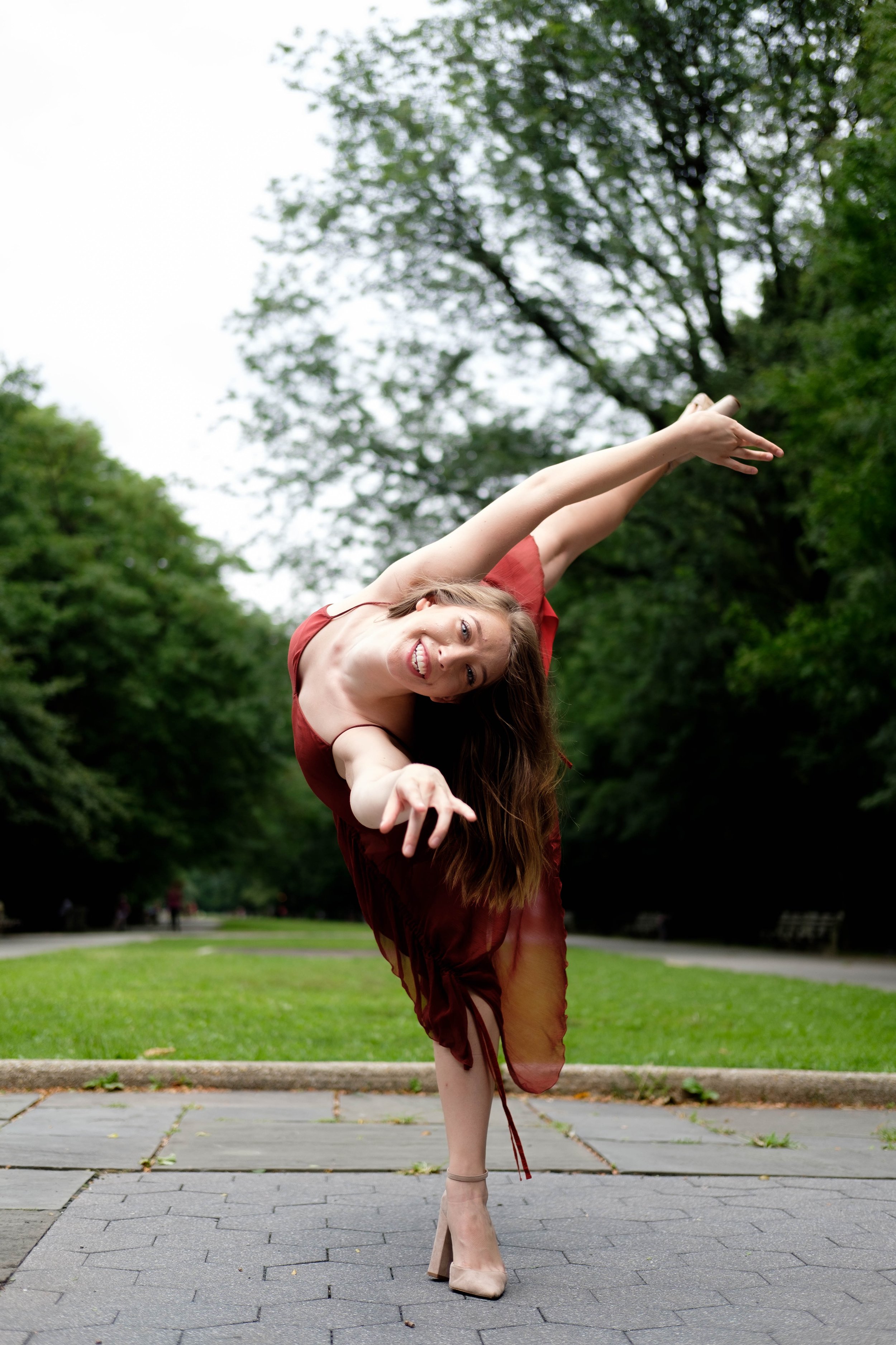 A woman with long hair dancing outdoors in a park, wearing a red dress and nude high heels, with trees and green grass in the background.