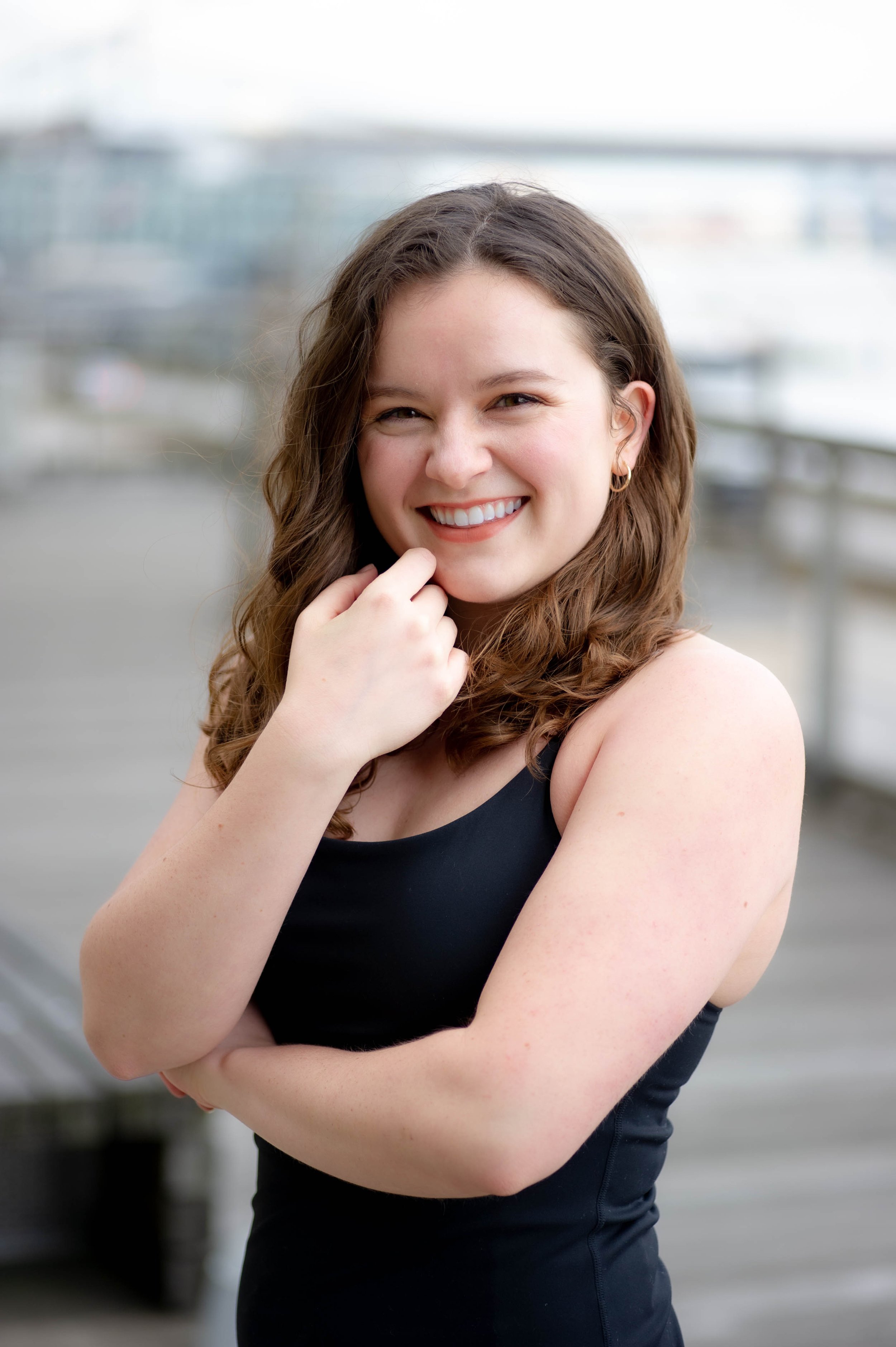 A young woman with long brown curly hair smiling outdoors, wearing a black sleeveless top, with cars and a waterfront in the background.