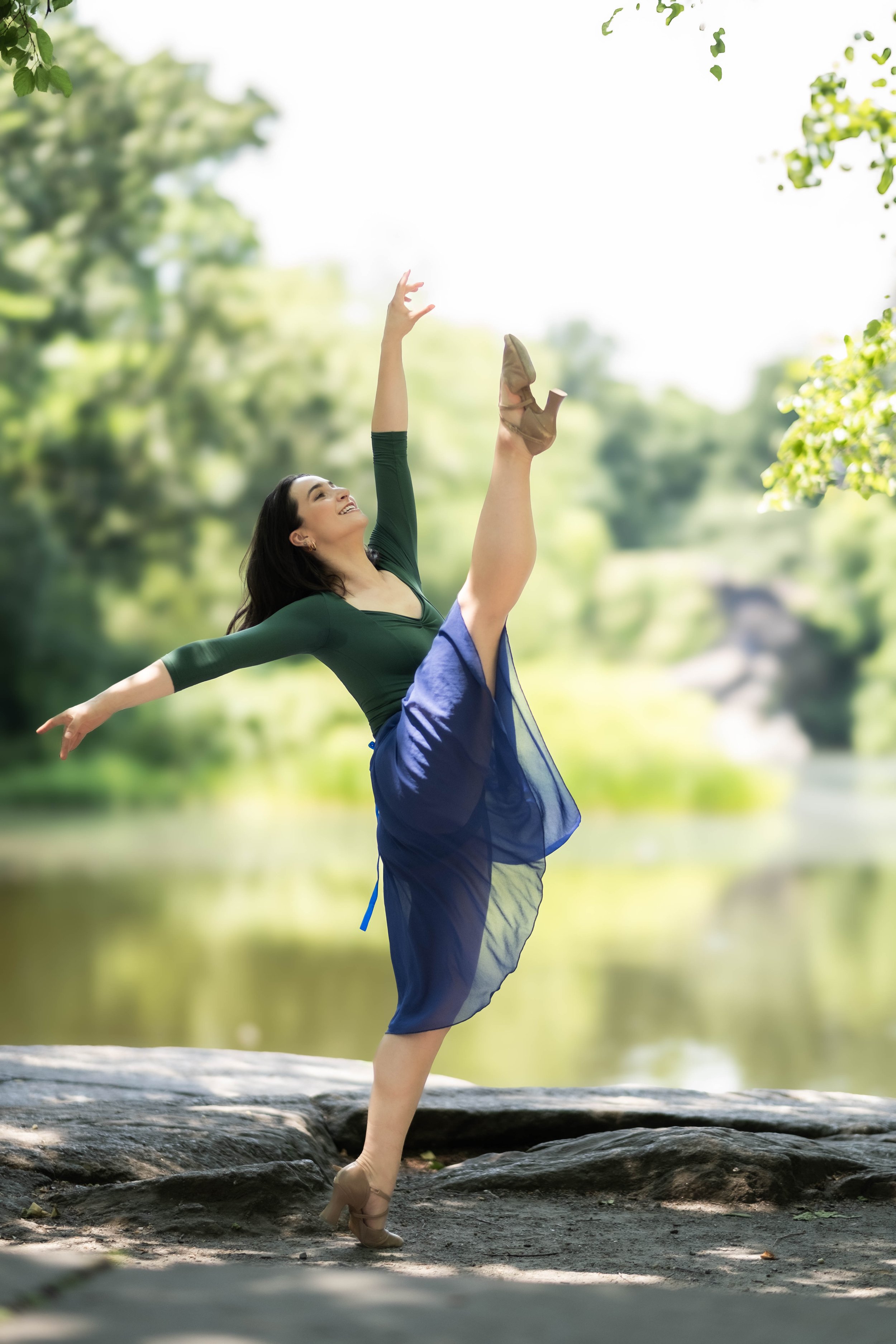 A woman ballet dancer wearing beige pointe shoes, a green long-sleeve top, and a blue tulle skirt, practicing ballet outdoors on a log near a body of water with trees in the background.