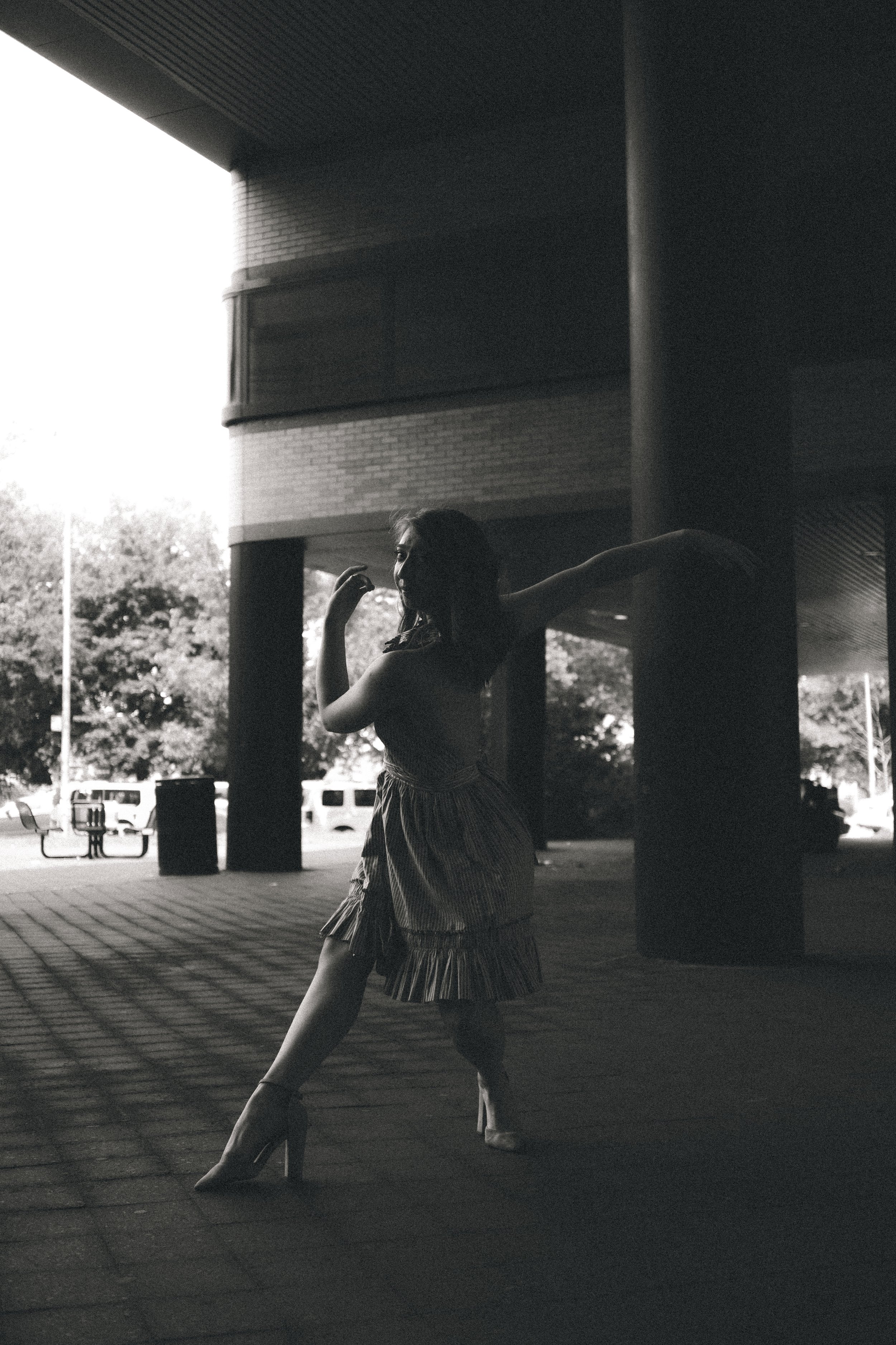 A silhouetted woman in a flowing skirt and high heels, posing with one arm bent near her face and the other extended outwards, standing under a building's overhang with trees and parked cars in the background.