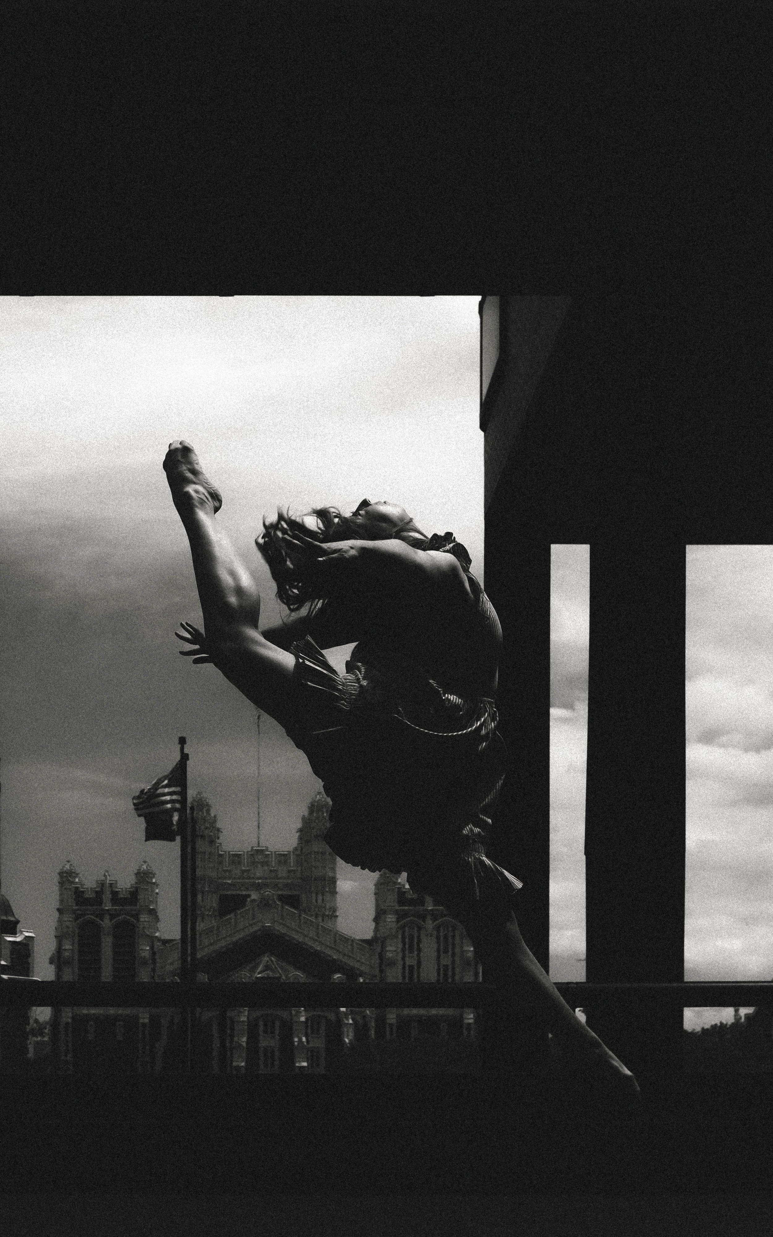 Black and white photo of a woman dancing or performing a pose outdoors with architecture and American flags in the background.