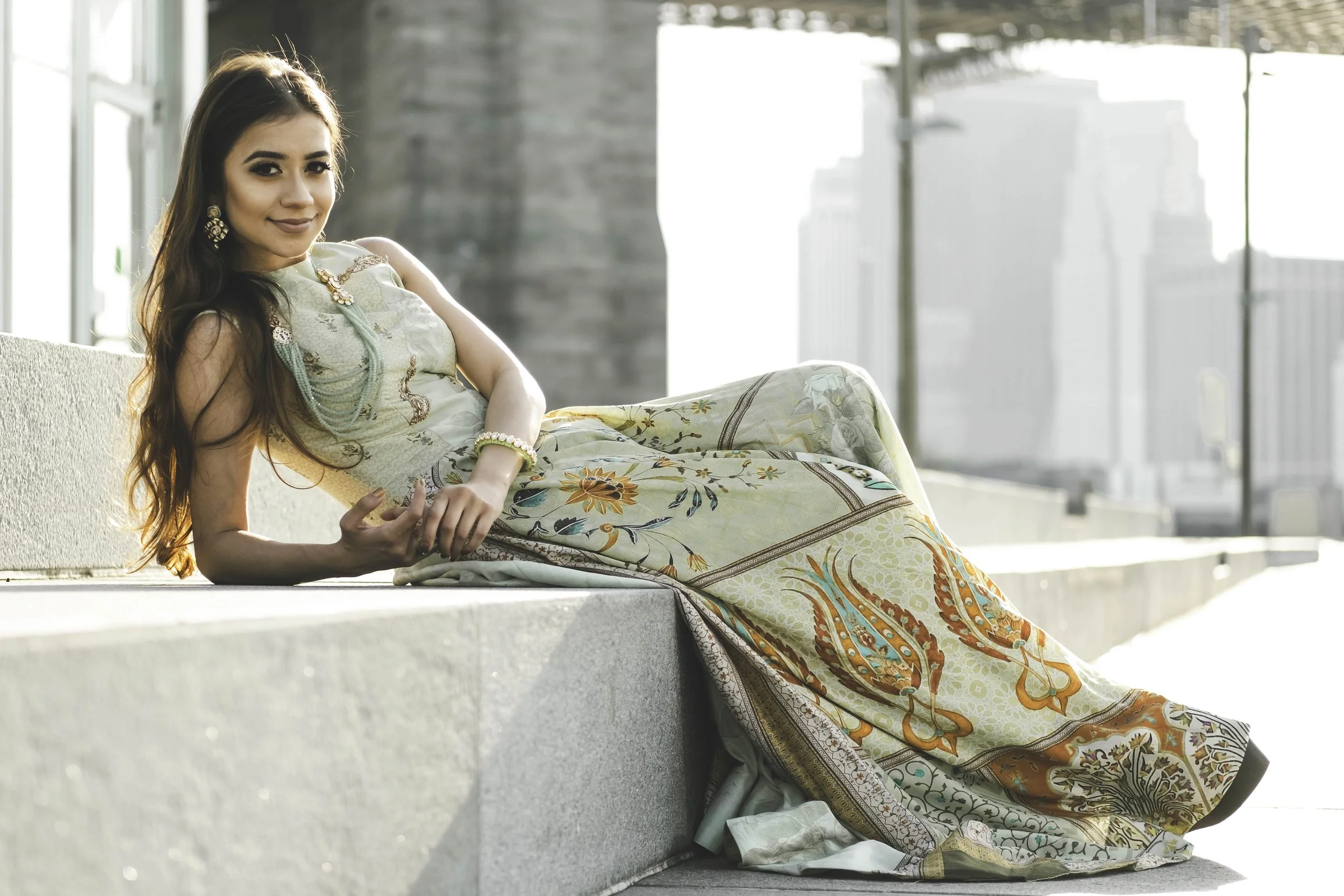 A woman with long dark hair sitting on an urban balcony ledge, wearing a colorful patterned dress and jewelry, smiling softly at the camera with city buildings in the blurred background.