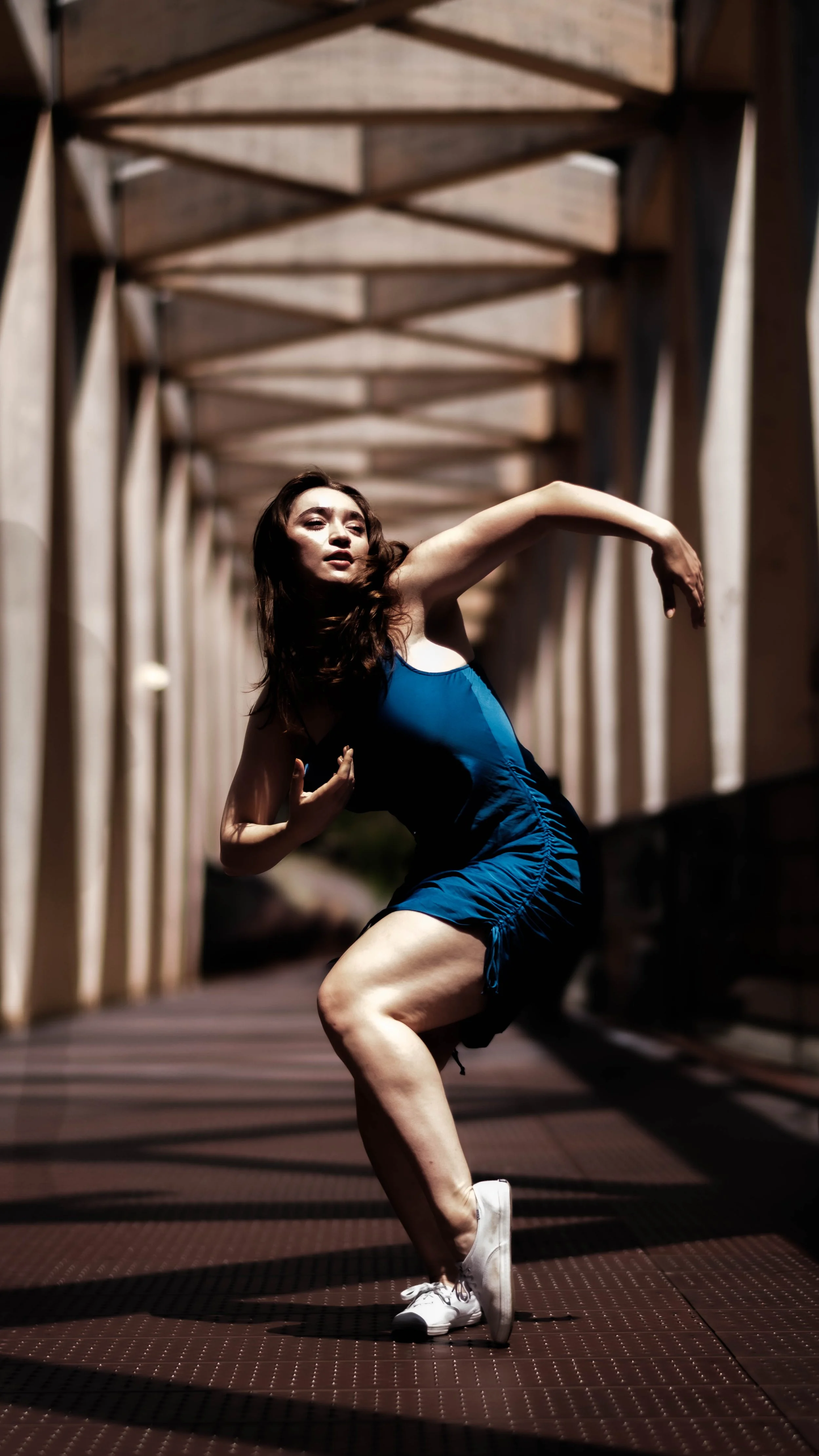 A woman in a blue dress and white sneakers dancing on a bridge with wooden beams overhead.