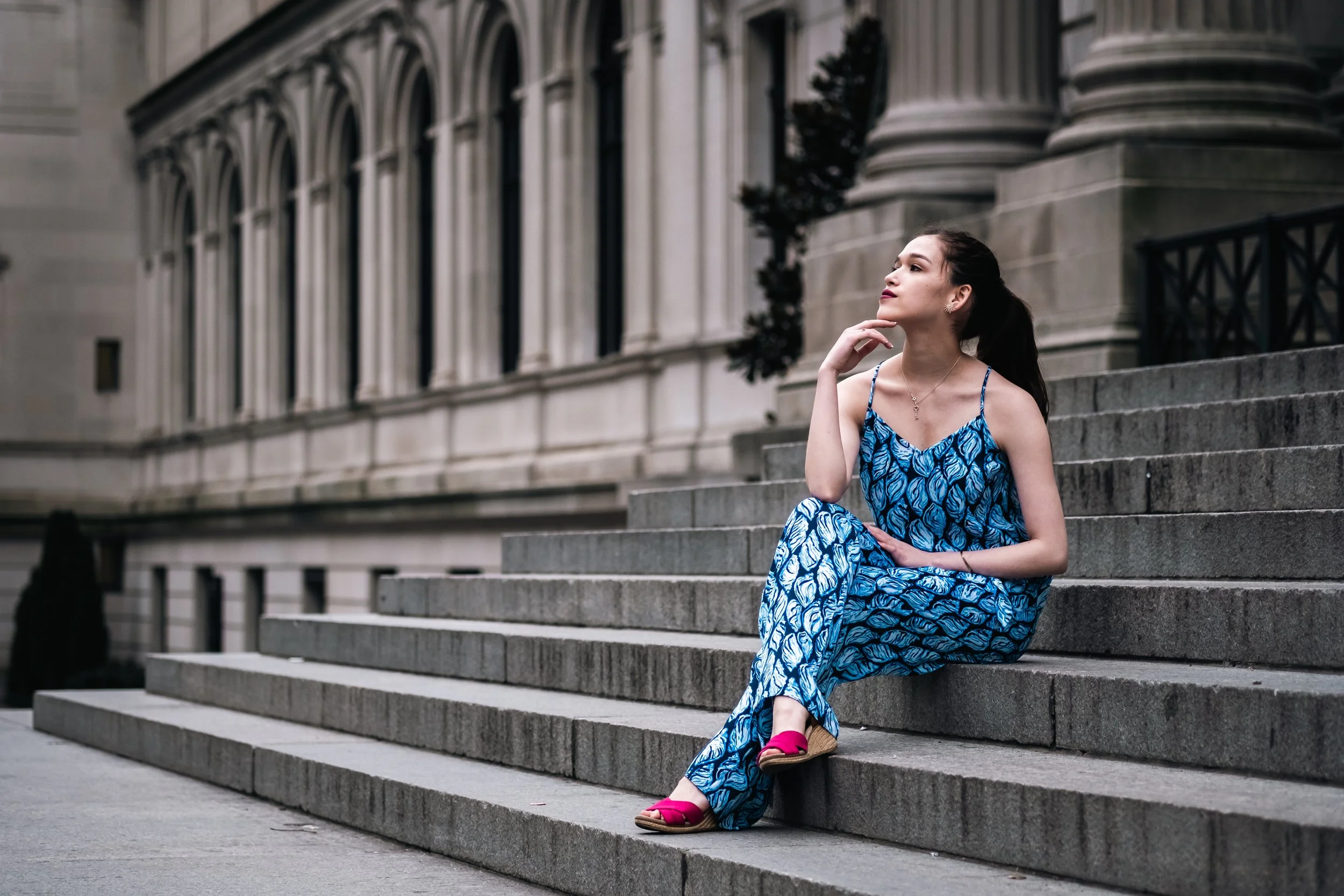 A young woman in a blue patterned dress and pink sandals is sitting on stone steps outside a historic building with large arched windows and ornate stonework.