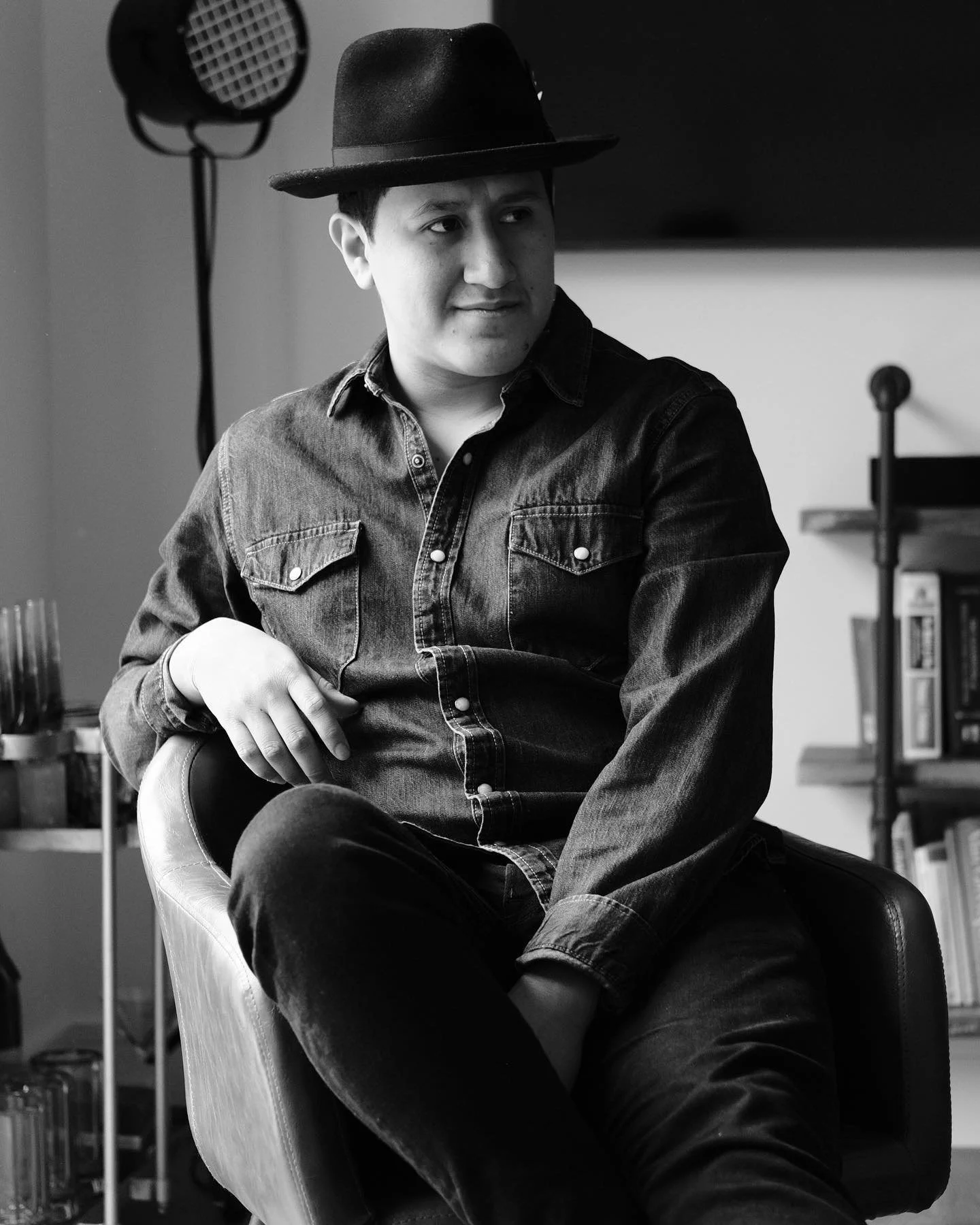 A black and white photo of a man sitting in a chair, wearing a dark hat and a denim button-up shirt, with a bookcase and studio light in the background.