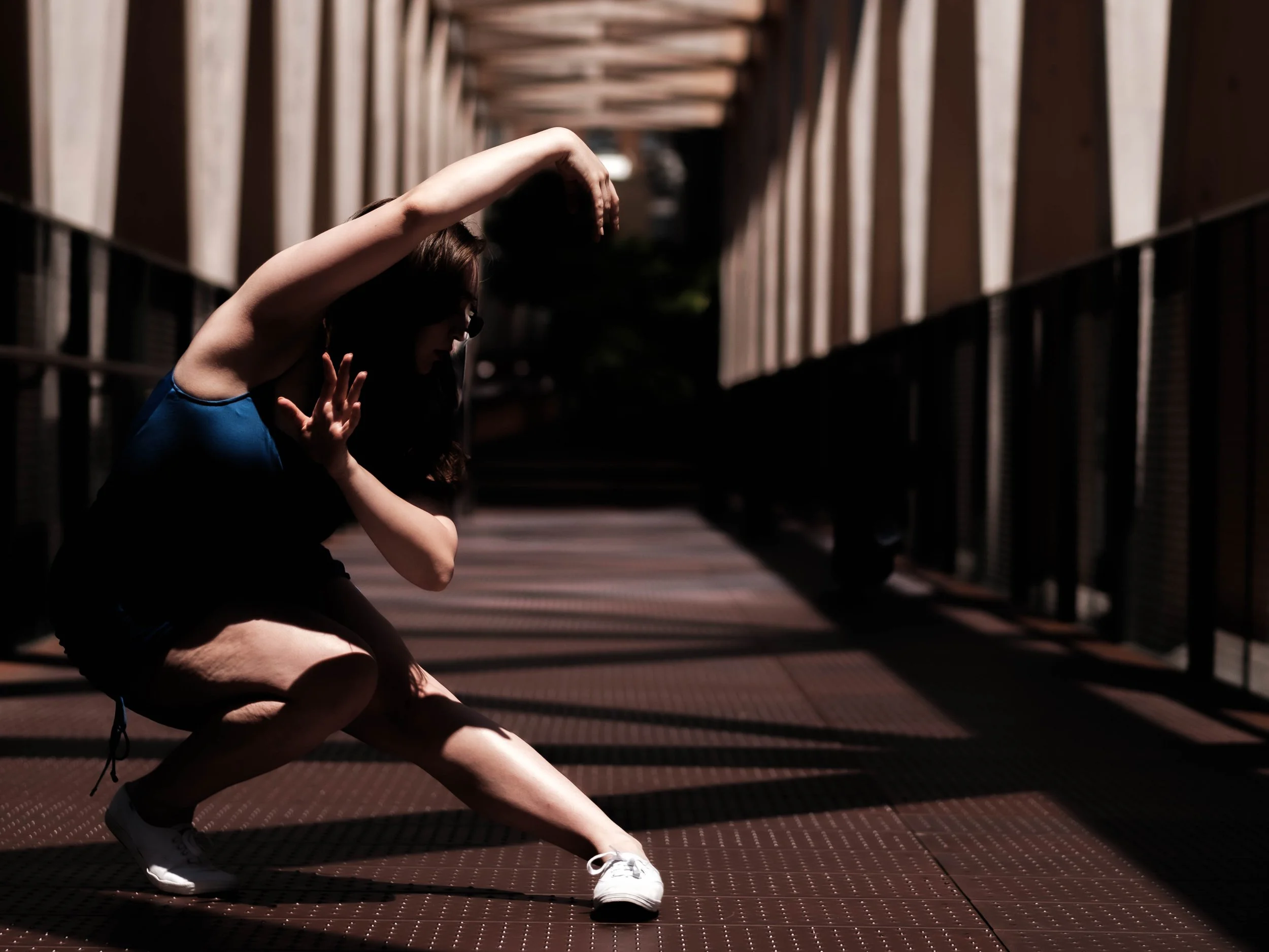 A woman in athletic clothing doing a stretching exercise outdoors on a bridge, with her left arm raised overhead and her right hand touching her face, in a shadowed setting with sunlight filtering through the structure.