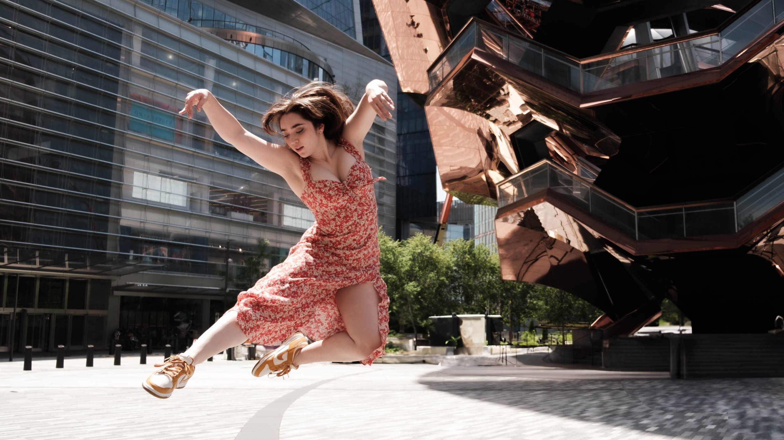 A woman in a red floral dress and sneakers is jumping in front of modern city buildings and a reflective, geometric sculpture.