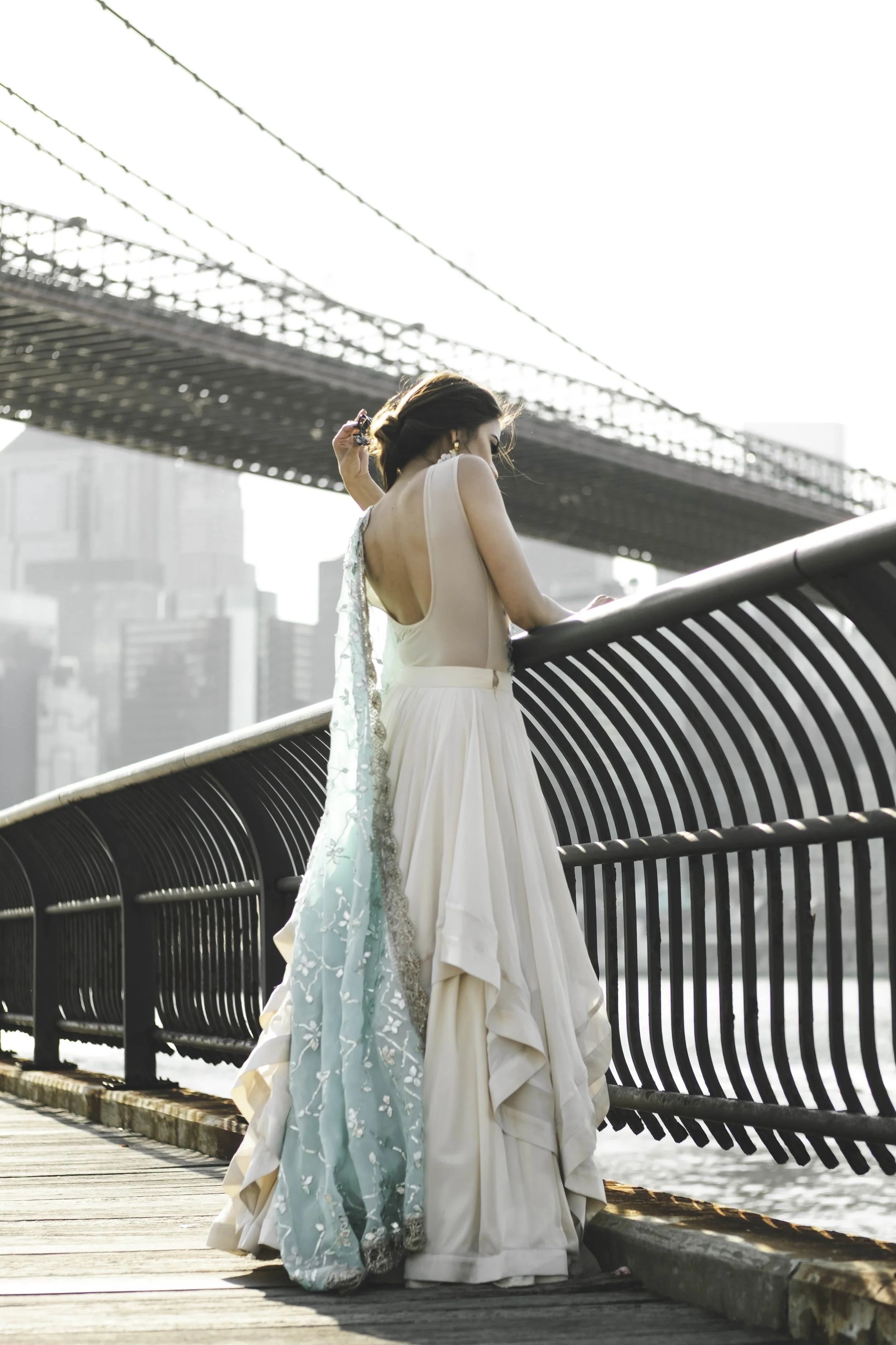 A woman in a flowing white and mint green dress standing on a bridge, looking down as the city skyline and a bridge are visible in the background.