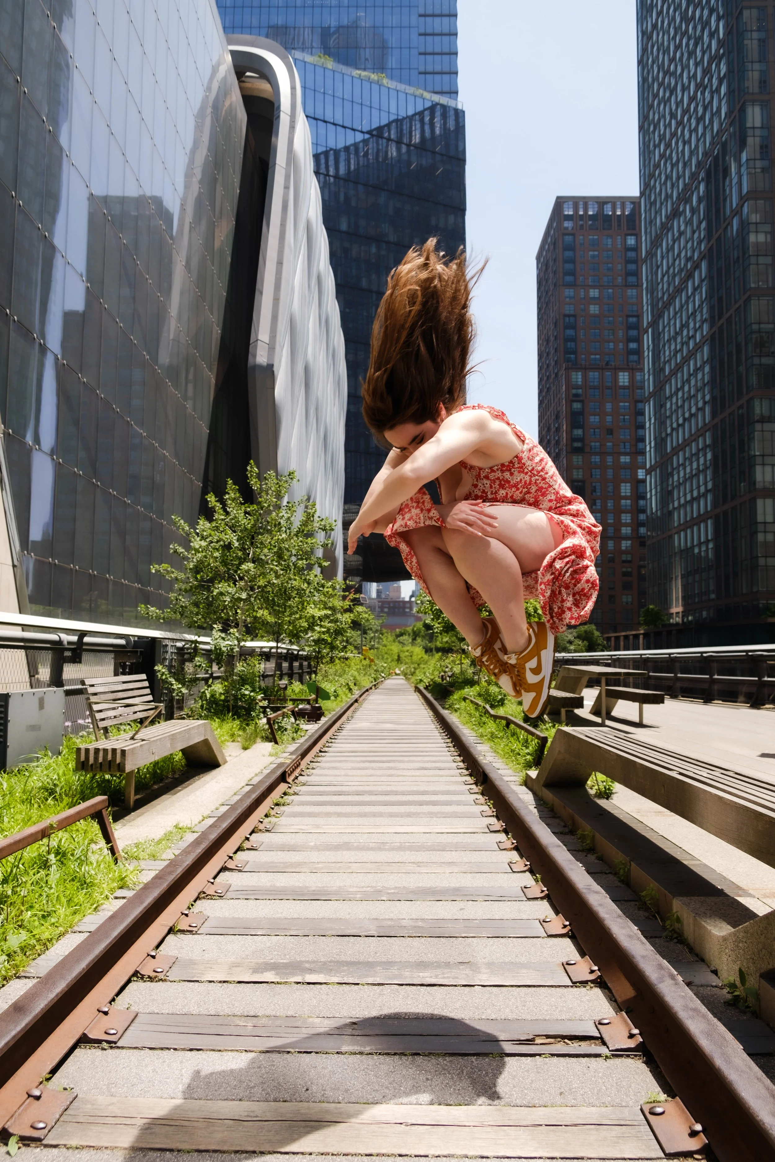 A woman with long hair jumping on railway tracks in an urban park surrounded by tall buildings and benches.