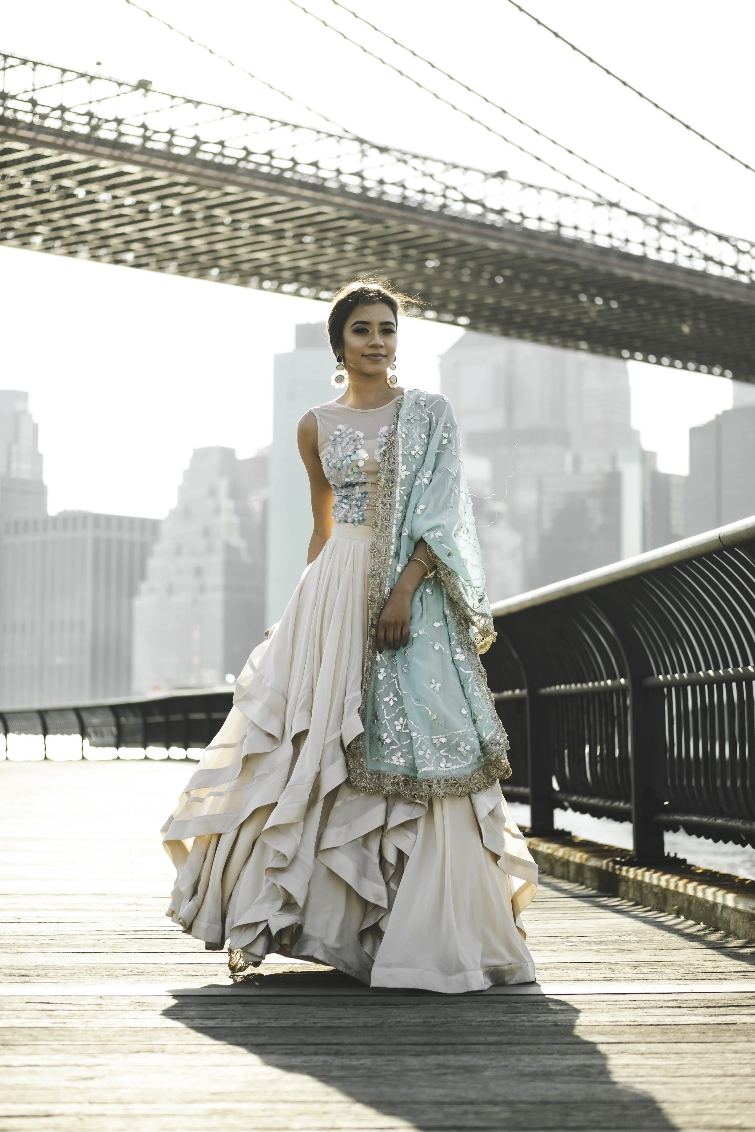 A woman wearing a traditional white and teal gown with embroidery, standing on a wooden boardwalk with city buildings and a suspension bridge in the background, during daylight.